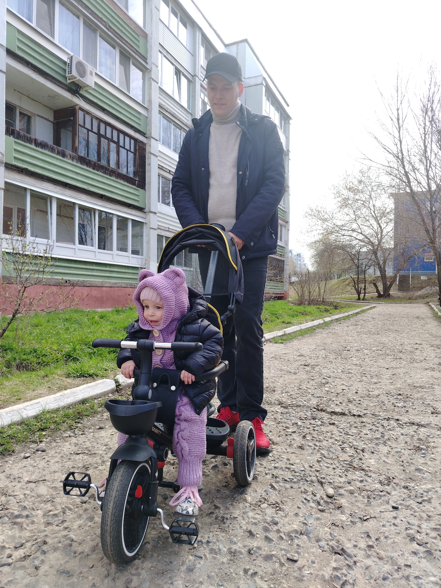 A man and a child are on a sidewalk in Chaikovsky, Russia. The man is wearing a dark-colored baseball cap, a navy-blue jacket, and black pants, with bright red shoes. He is standing behind a child's tricycle, likely pushing it. The child is wearing a dark puffy jacket and a full purple knitted outfit, including a hat with ears. The tricycle is black with a small storage bin in the front. The sidewalk is paved with gravel and pebbles, and there are apartment buildings in the background with bare trees nearby. There are curb-stones on both sides of the sidewalk.