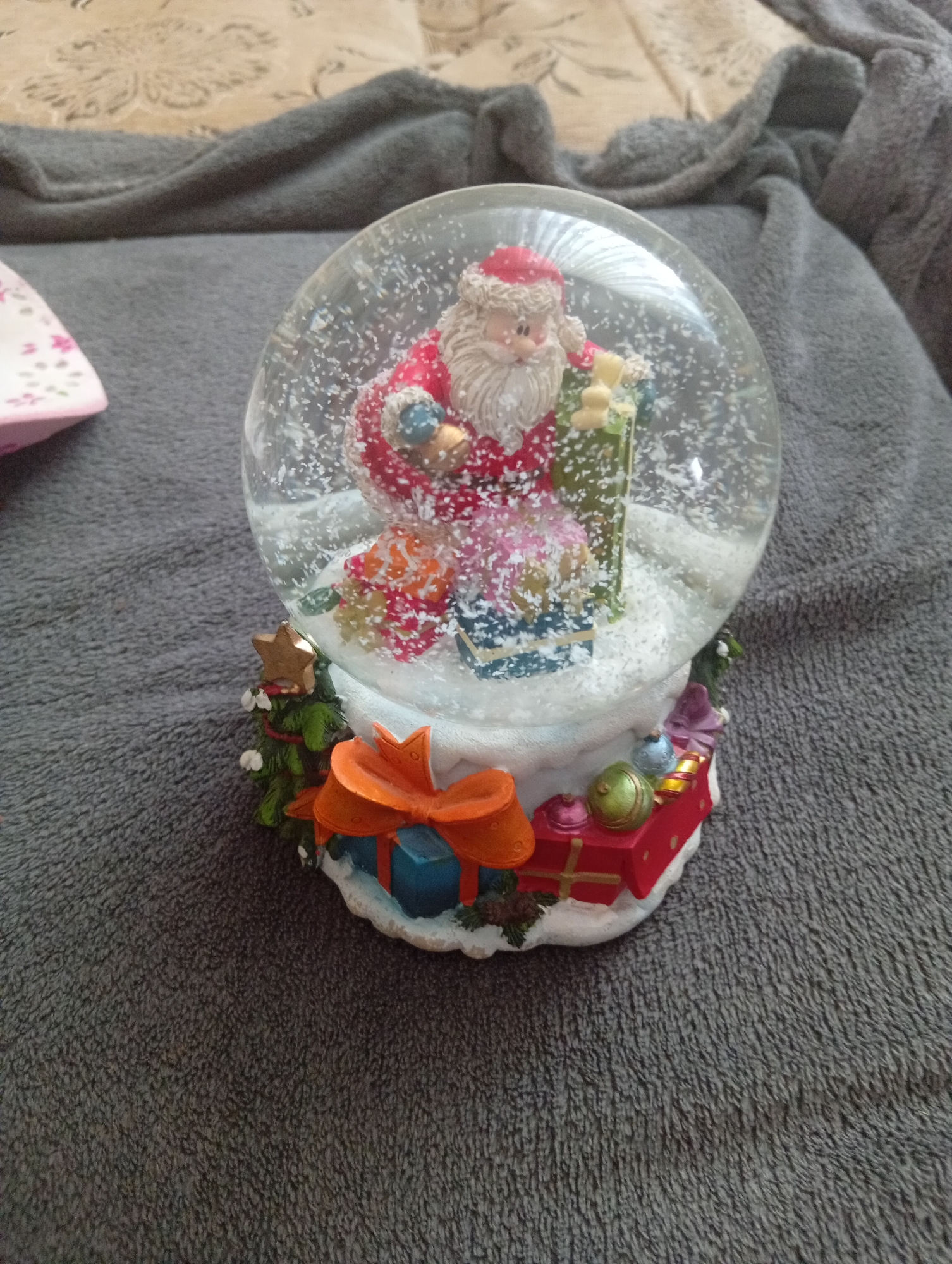 This is a close-up, eye-level photograph of a Christmas-themed snow globe. The snow globe is centrally located, with its clear glass dome filled with artificial snow and a miniature scene. Inside, a figure of Santa Claus, depicted with a white beard, red hat, and red suit trimmed with white fur, stands amongst presents and a small Christmas tree. The base of the snow globe is decorated with miniature festive elements, including pine branches, a star, and several wrapped gifts, one of which is topped with an orange bow. The snow globe rests on a textured, dark gray surface, likely a blanket or sofa, with a portion of a beige, patterned fabric visible in the upper background. The overall impression is one of a festive indoor display. No information is available to confirm the location of Veliatyno, Ukraine.
