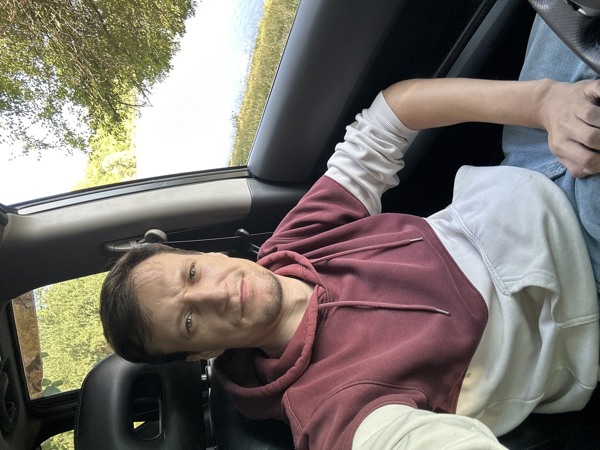 A medium shot captures a man sitting in the driver's seat of a car, with a body of water and trees visible through the passenger-side window. The man, with short brown hair and a light beard, is wearing a maroon and cream-colored hoodie. His right arm rests on his lap, and his left arm is slightly bent. The car's interior is dark, contrasting with the bright, sunlit exterior. The scene is not identifiable as being in Makiv, Ukraine, or any specific city.