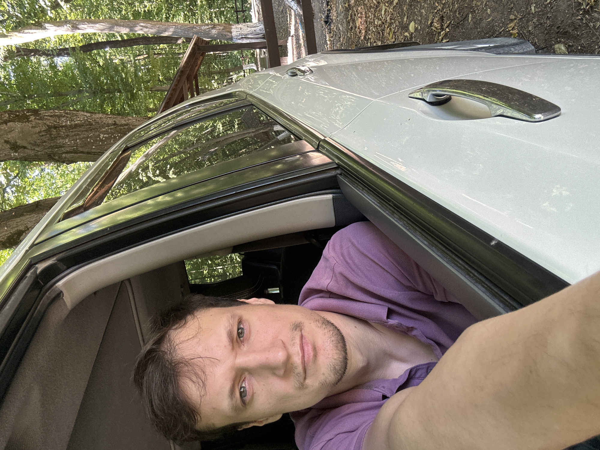 A medium shot photograph shows a man sitting in the driver's seat of a white car. The man has light skin, light brown hair, and a light beard. He is wearing a purple collared shirt and is looking directly at the camera. His left arm is resting on the open car door, obscuring part of his torso. The interior of the car, including the headliner and the dashboard, is visible. The car is parked in a wooded area, with trees and foliage visible through the open car door and windows. A wooden picnic shelter and a wooden fence are also visible in the background. The ground is covered with fallen leaves and dirt. The overall lighting suggests it is daytime. The location is in the city of Shatava, Ukraine.