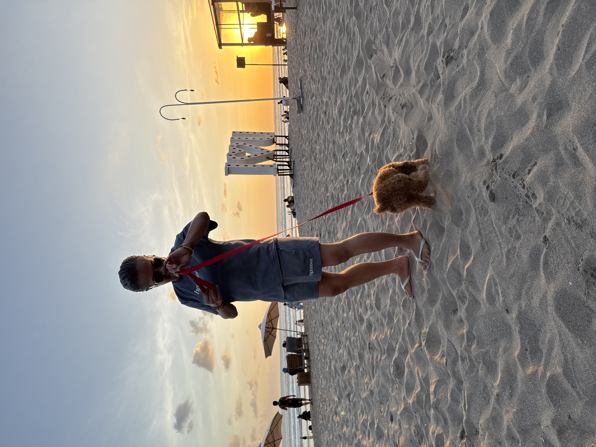 At sunset on Kuta Beach, Bali, Indonesia, a person wearing sunglasses, a grey t-shirt, and grey shorts stands on the sand, holding a red leash attached to a small, brown, curly-haired dog. The person is looking down at their phone. In the background, several large white letter sculptures spell out "I LOVE BALI". Beach umbrellas and chairs are visible, as are other beachgoers. The sky is a gradient of blue and orange, indicative of a sunset. The sand is rippled by the wind.
