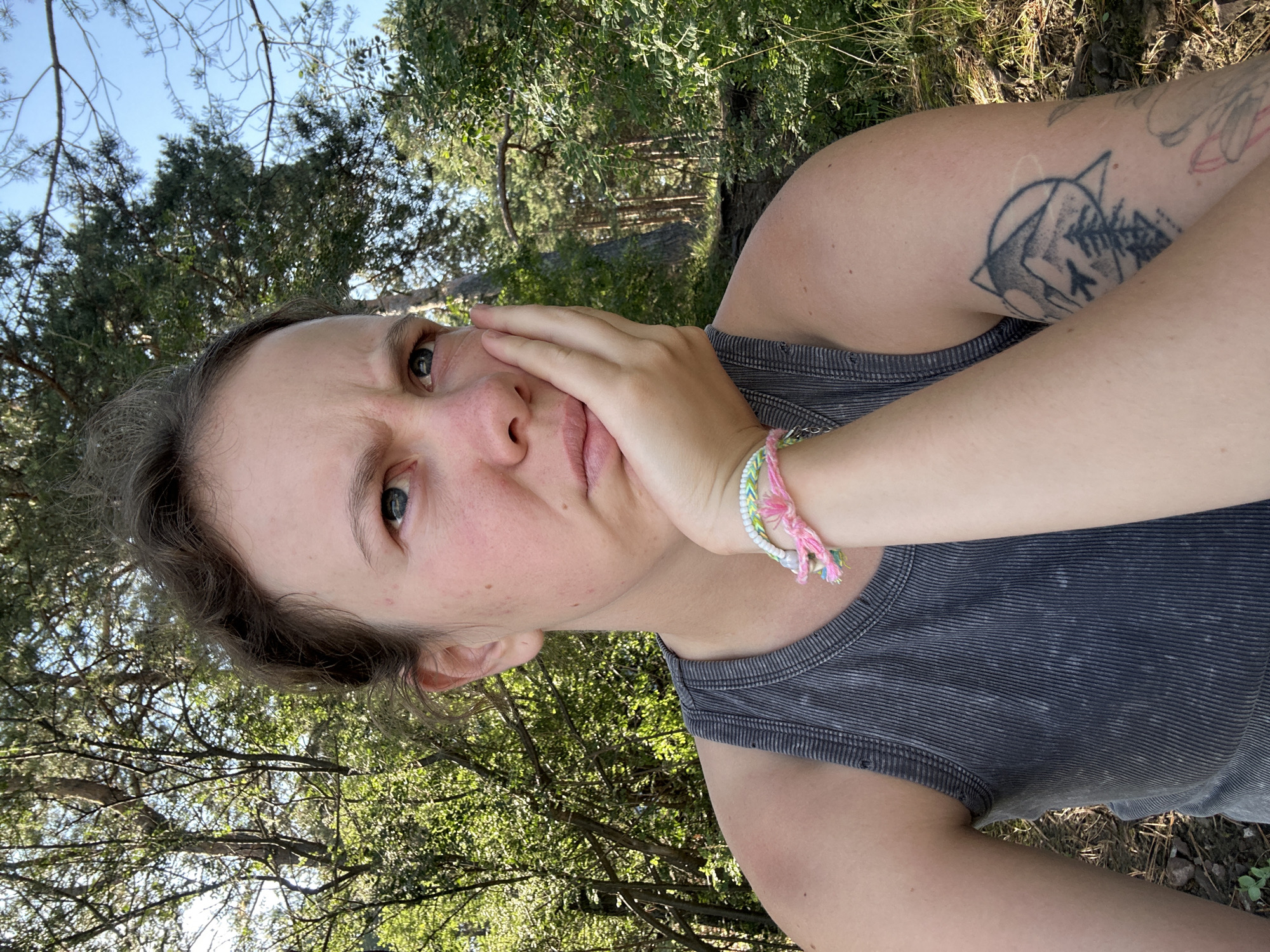 A young woman with her hand on her cheek is looking off to the side. She is wearing a grey tank top and has a bracelet on her wrist. She has a tattoo on her arm. The background is out of focus and appears to be a forest with trees and sunlight. There is no indication of the city of Nikolo-Petrovka, Russia in the image.