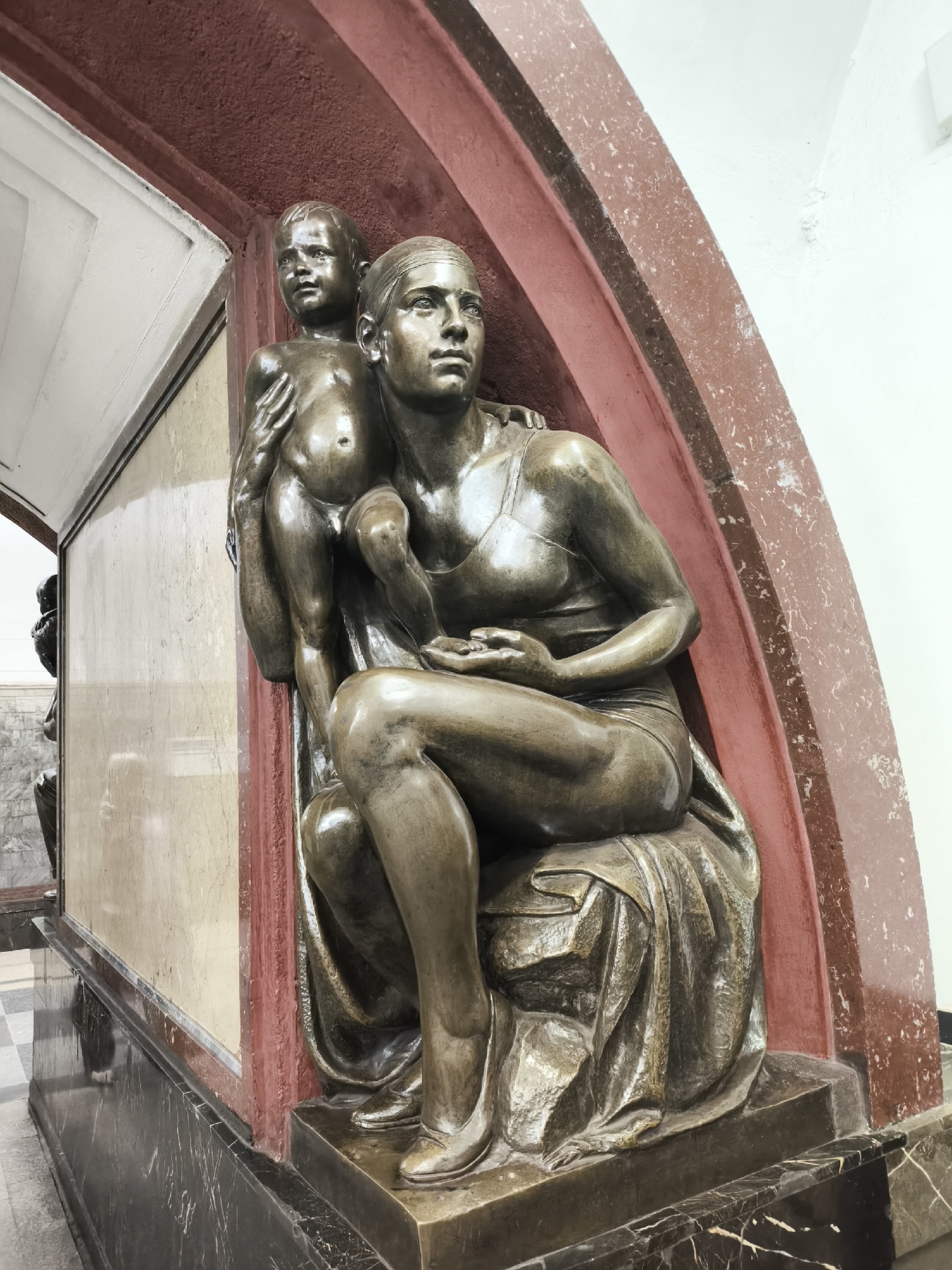 A low-angle, full shot shows a sculpture of a woman holding a small child in a subway station. The statue is made of bronze-colored metal, and appears to be situated in a alcove that has a red archway. The woman is seated, cross-legged, wearing a bathing suit and a serious expression. The child, naked and clinging to the woman's arm, looks on with a similar serious look. The background features light-colored, veined marble and other sculptures further back along the wall. The location is an unknown subway station in an unknown city.