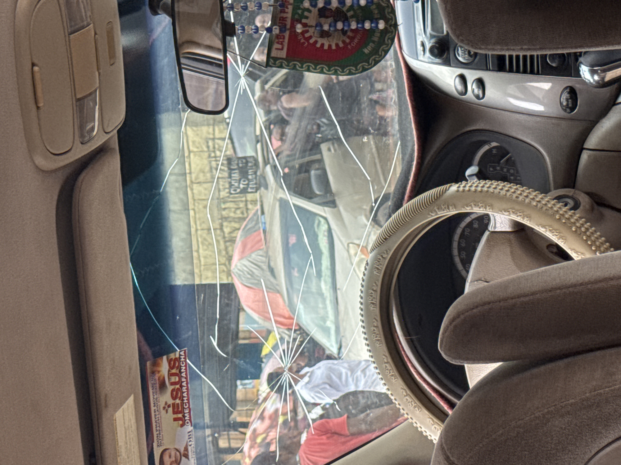 The image, taken from inside a vehicle, shows a cracked windshield obscuring a street scene in Orji, Nigeria. Through the damage, several people are visible, some standing and others near or inside vehicles. One person in the foreground, inside the car, has their arms resting on the steering wheel. Outside the car, there are at least two individuals dressed in casual attire, with one wearing a red t-shirt and the other a white shirt. Further in the background, more people and vehicles are present, with umbrellas providing shade in what appears to be a marketplace or busy street. A political sticker with the words "LABOUR PARTY" is hanging from the rearview mirror. The overall environment suggests a busy urban setting, likely during the daytime.