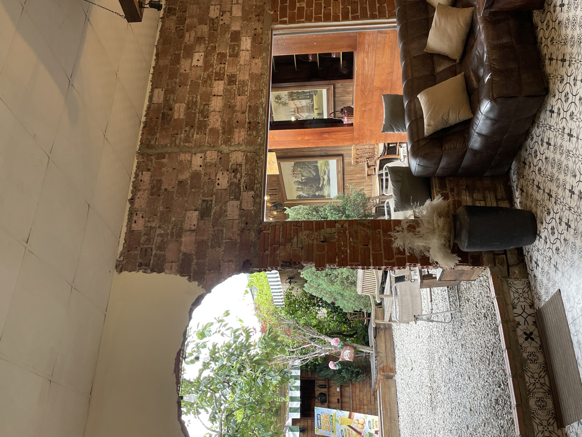 The image depicts an indoor/outdoor cafe space in Phường Xuân Hương, Đà Lạt, Vietnam. The foreground is dominated by a dark brown leather sofa with beige and grey cushions, situated on a patterned tile floor. To the left, a large, rough-hewn opening in a brick wall reveals an outdoor courtyard. This courtyard is filled with gravel and features a variety of elements including trees, foliage, a wooden bench, a metal chair, a wooden planter box, and a promotional sign for "COMBO CHI 55K" with images of drinks. In the background of the courtyard, there are striped awnings and more greenery. Further inside the cafe, through another opening in the brick wall, framed paintings of buildings and landscapes are visible, along with wooden shelves and decorative baskets. A dark grey vase with tall, dried reeds stands next to the sofa, adding to the rustic ambiance. The overall impression is of a cozy, eclectic cafe with a blend of indoor comfort and outdoor relaxation.