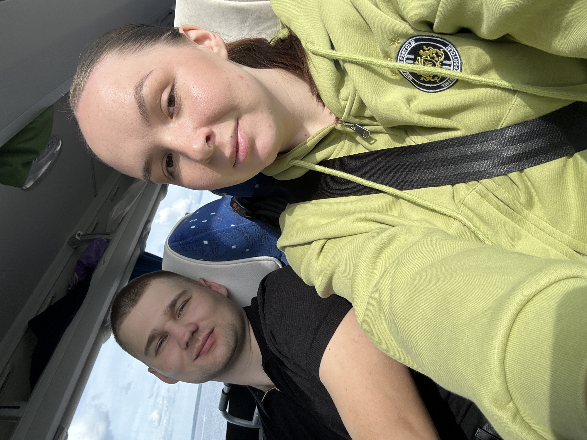 A selfie captures two young individuals, a woman and a man, seated within what appears to be a bus or train, presumably traveling in Chaikovsky, Russia. The woman, in the foreground, is taking the photo, looking directly at the camera with a neutral, slightly smiling expression. Her dark hair is pulled back, and she is wearing a vibrant lime green hooded sweatshirt. A prominent black seatbelt crosses diagonally over her chest.
On the woman's sweatshirt, a circular emblem is visible on the left chest area, featuring a gold and black design with text. Partially legible text on the emblem includes "FSK CLM" at the top and "199 EG" at the bottom.
Behind the woman, a young man is also visible, looking towards the camera with a relaxed expression. He has short, light brown hair and is wearing a black t-shirt. The seat behind him features blue and white patterned upholstery. Through a window to the left of the man, a bright, partially cloudy sky is visible, indicating that the photo was taken during the daytime. The interior of the vehicle includes overhead compartments, characteristic of public transportation. The scene depicts a casual journey or commute.