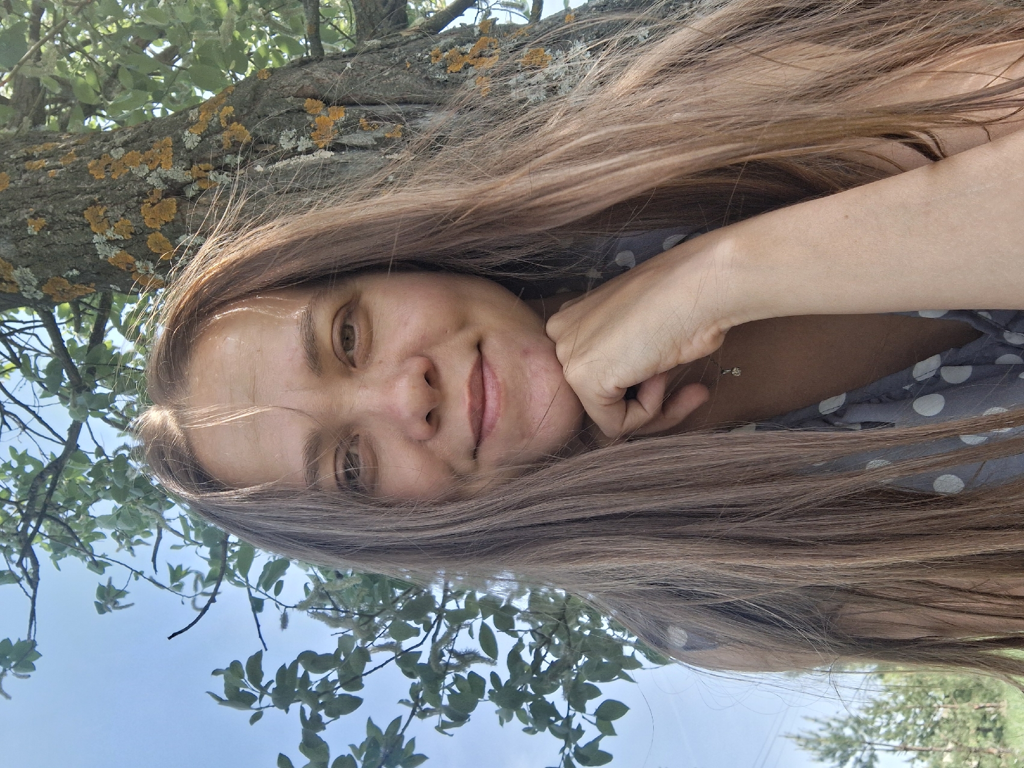 A young woman with long brown hair rests her chin on her hand while lying on a tree branch. She is wearing a grey polka-dot shirt. The bark of the tree is visible, covered with moss and lichen. The background consists of blue sky and green foliage. This image was taken in the city of Chaikovsky, Russia.
