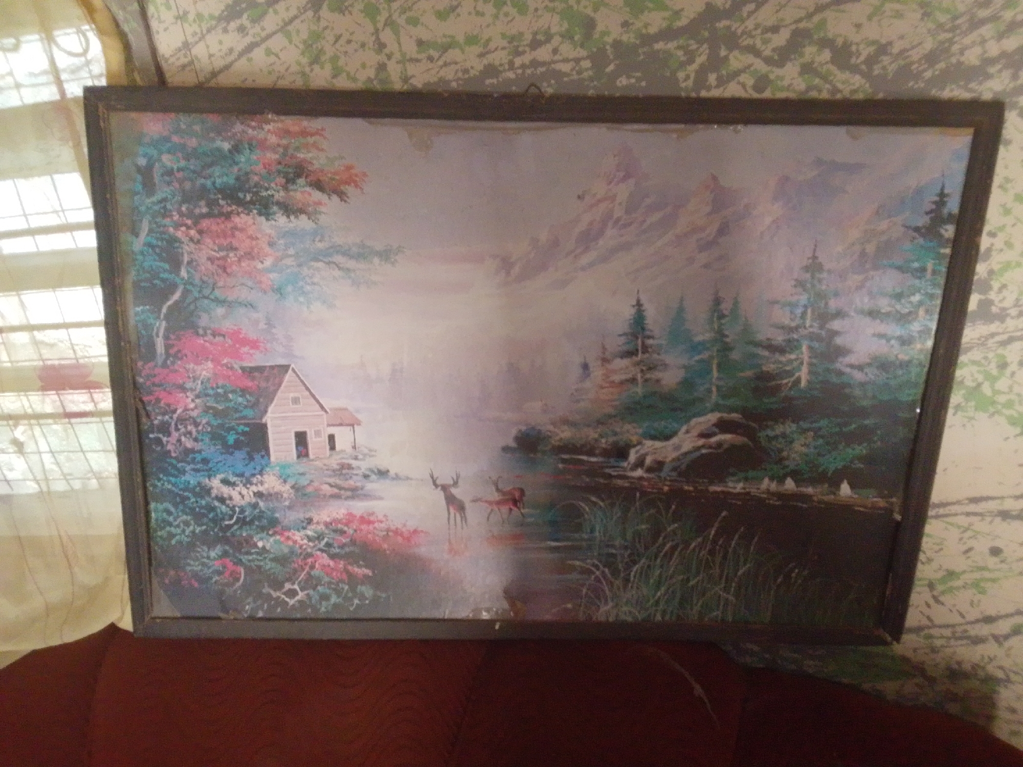 The image is a framed painting of a serene landscape scene. The painting depicts a rustic house nestled amongst vibrant trees with pink and blue foliage. In the foreground, a body of water reflects the soft, misty sky and distant mountains. Two deer are visible in the water, one standing alert and the other grazing. The painting is hung on a wall with a textured wallpaper featuring a pattern of green and grey splotches. To the left of the painting, a sheer curtain with a grid pattern is visible, suggesting a window. The bottom of the image shows a portion of a dark red upholstered surface, likely furniture. The overall impression is one of tranquility and natural beauty. The provided context states the location is Jalingo, Nigeria.