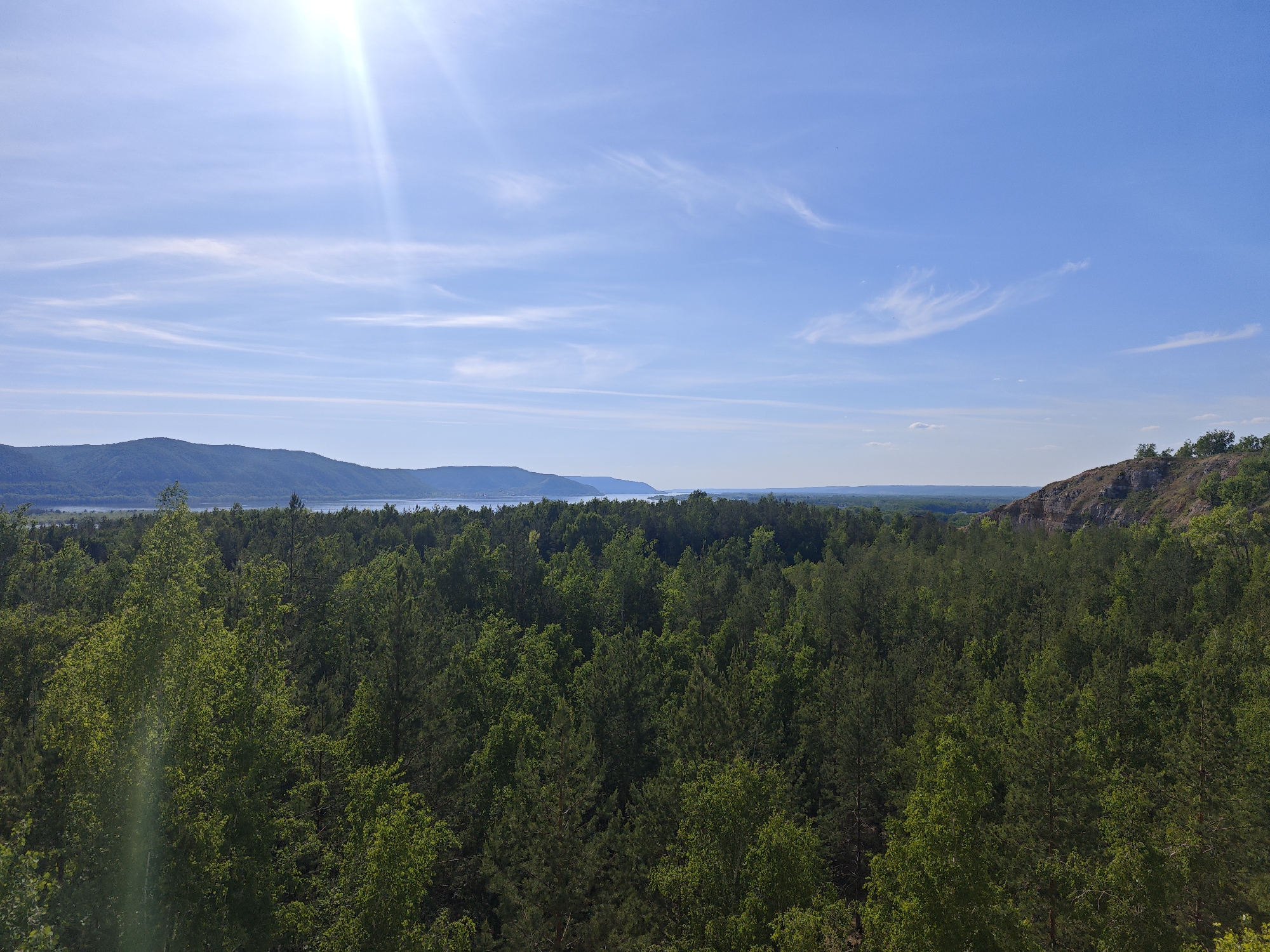 The image depicts a landscape view on a bright, sunny day with a clear blue sky and some wispy clouds. Sunlight is visible at the top left. In the background, there are mountains or hills partially obscured by a body of water, possibly a river or lake. The foreground is dominated by a dense forest with various shades of green foliage. To the right, a rocky cliff or outcrop is present, with some vegetation growing on it. The location is possibly near the city of Volzhskiy, Russia, based on the presence of hills and forests near a body of water.