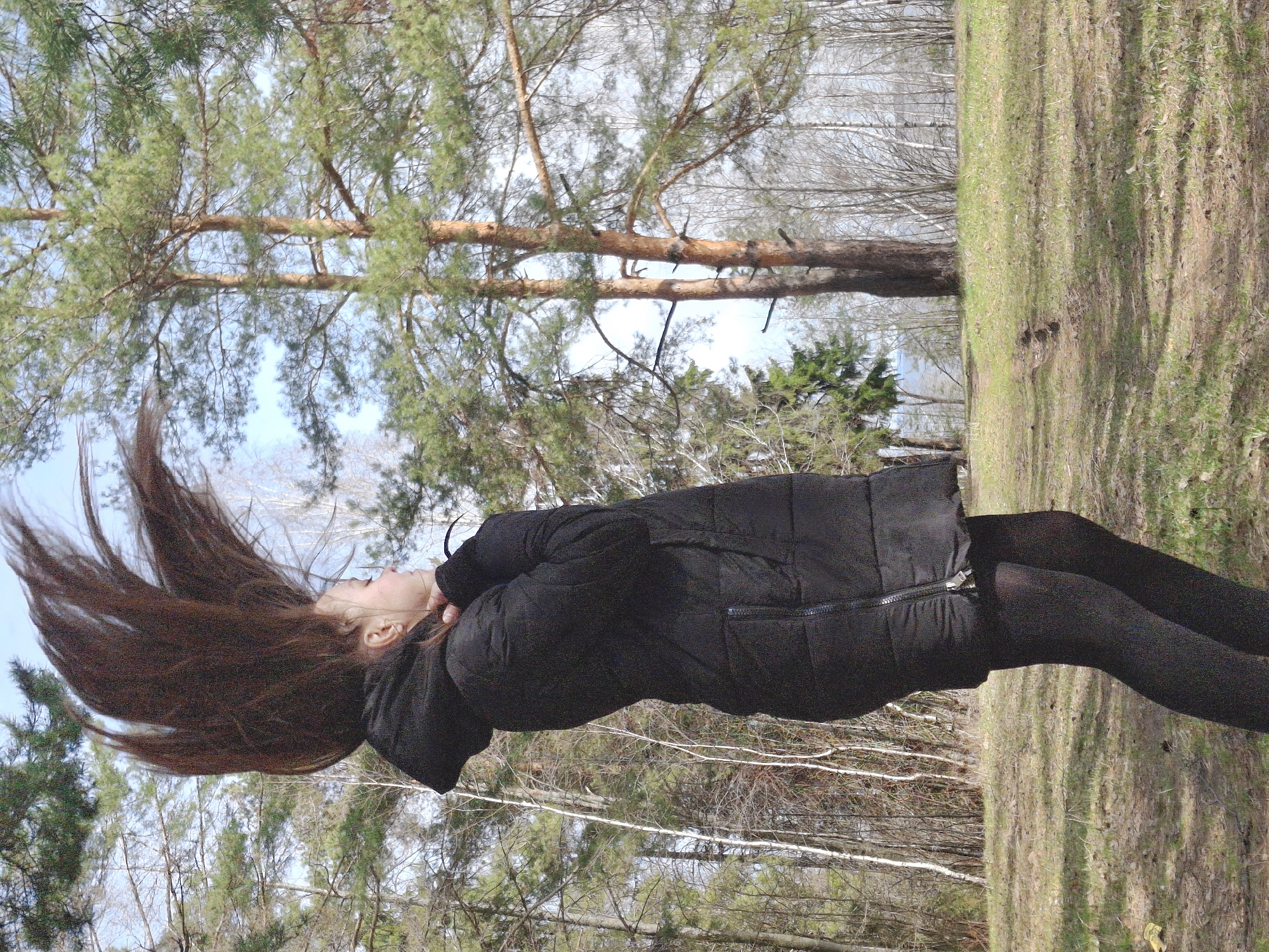 A young person with long, dark brown hair is captured in a dynamic pose, their head tilted back and hair flung upwards, suggesting movement or a dramatic gesture. They are wearing a black, padded winter coat with a zipper closure and dark leggings or tights. The subject is standing on what appears to be a grassy or mossy forest floor, which has patches of dry brown leaves and some green growth.
The background is a natural outdoor setting, likely a forest or park in Chaikovsky, Russia. Tall pine trees with green needles and light brown trunks dominate the upper part of the image, while the mid-ground and lower background feature deciduous trees, some of which are bare-branched, indicating late winter or early spring. The sky, visible through the tree canopy, is light blue with some haziness, suggesting daylight. The lighting is natural, casting some shadows on the ground. The image is oriented vertically, with the person positioned on the right side and the forest floor appearing as a vertical strip on the right, while the trees extend across the rest of the frame. There is no visible text.