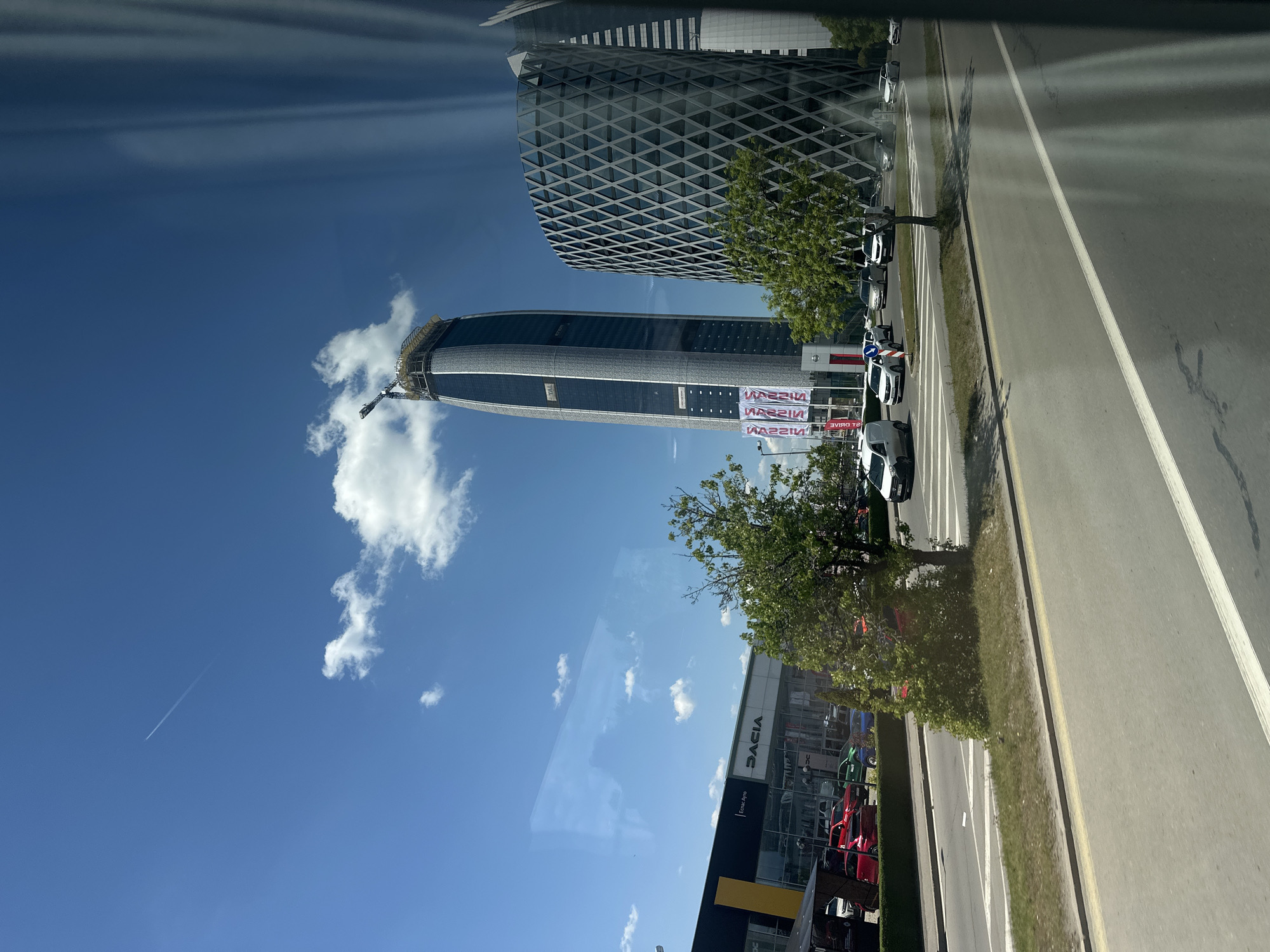 The image shows a view from inside a vehicle, possibly a bus, looking out at a street in Sofia, Bulgaria. On the right side of the road, there is a Dacia car dealership. A row of cars is parked alongside the road. Beyond the cars, there are two prominent office buildings. One is a tall, rounded skyscraper appearing to be under construction, with a crane visible at the top. The second building has a grid-like patterned facade. Banners with white text on a red background are visible near the skyscraper. The sky is blue with scattered clouds. A contrail from an airplane is also visible. The road is divided with painted white lines.