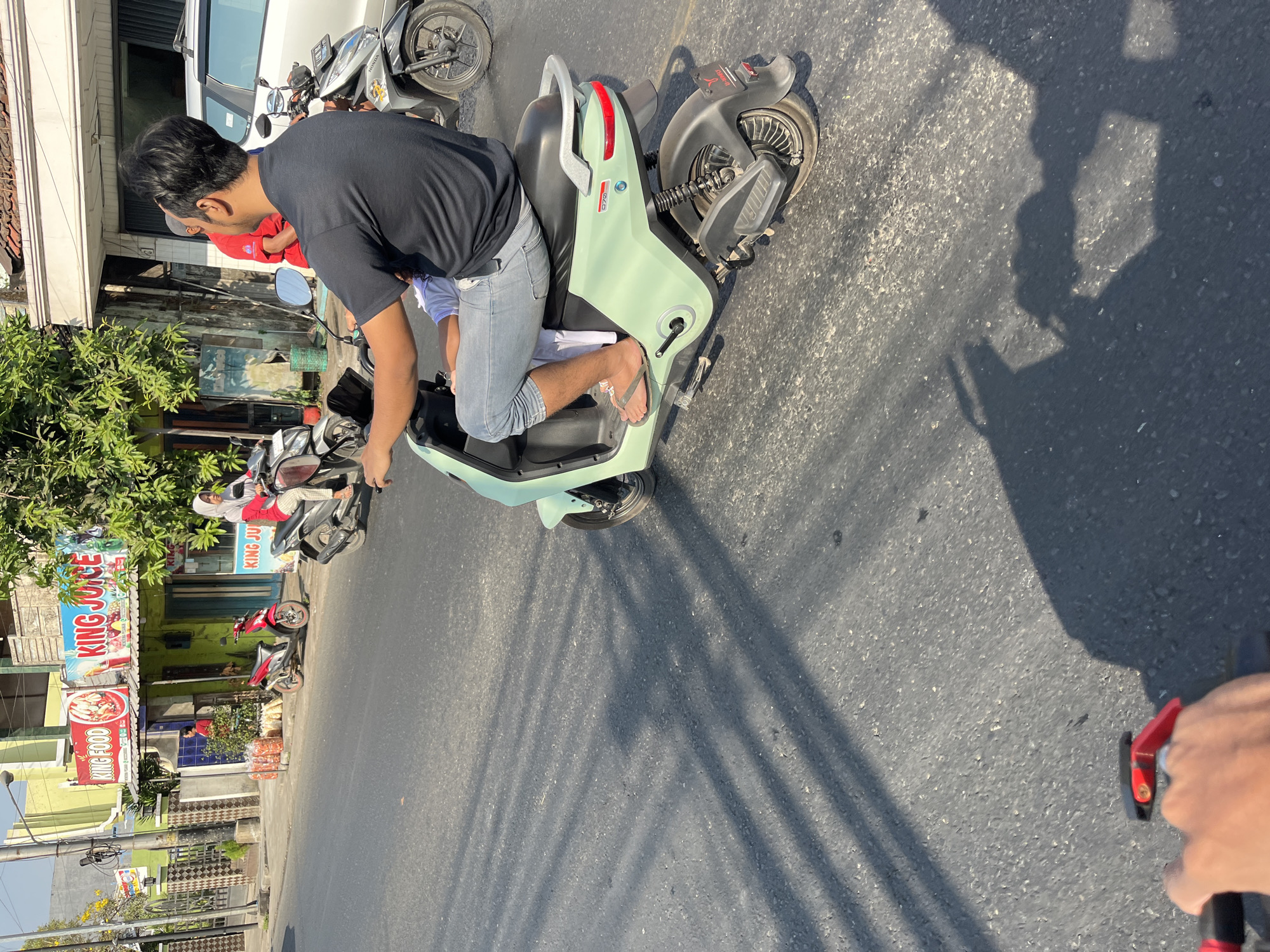 The image shows a street scene in Kalinyamatan, Indonesia, where a person is riding a light green electric scooter. The rider is wearing a black t-shirt and light blue jeans, with their legs dangling over the side of the scooter, wearing flip-flops. A child, wearing a white shirt, is seated in front of the rider. The scooter has a silver grab handle at the rear and a red taillight. In the background, there are several buildings, including one with prominent signs for "KING FOOD" and "KING JUICE." A red motorcycle is parked on the side of the road in front of one of the buildings. Other motorcycles and a white vehicle are also visible further down the street. Shadows from overhead structures and trees cast long lines across the asphalt road. The overall scene depicts a typical day on a street in Kalinyamatan.