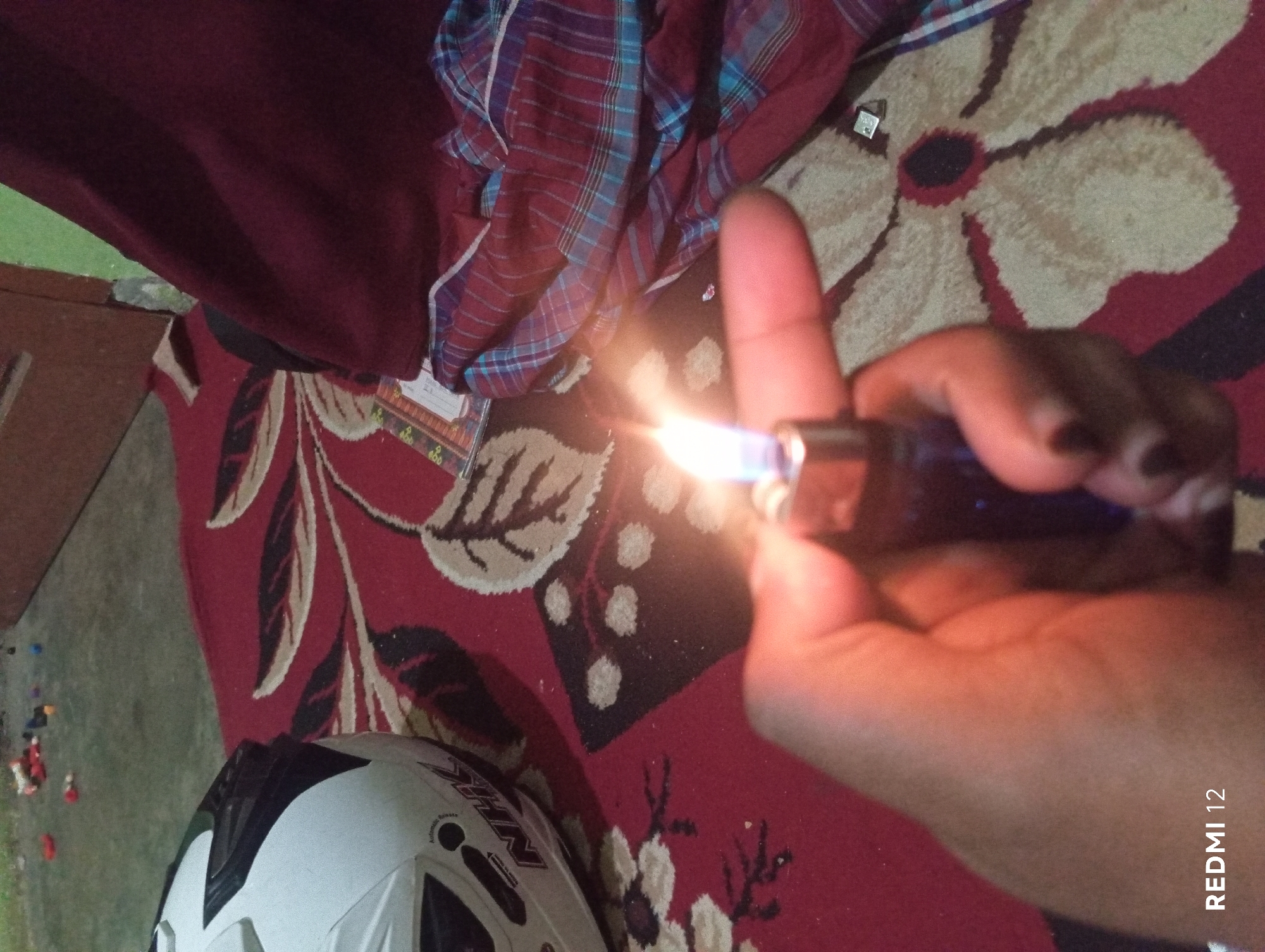The image captures a close-up view of a hand holding a lit blue lighter, with the flame emitting a bright yellow and blue light. The hand, with dark nail polish, is positioned in the foreground, obscuring part of the scene. In the background, a red rug with a floral pattern is visible. To the left, a white helmet with black accents is placed on the rug. Further back, a folded maroon and blue plaid fabric lies on the rug. In the upper left corner, a collection of small, colorful objects, possibly toys, are scattered on a grey surface. The overall setting appears to be an indoor environment, likely a room. The bottom left corner of the image displays the text "REDMI 12". There is no visual information that specifically indicates the location in the city of Pagar Alam, Indonesia.