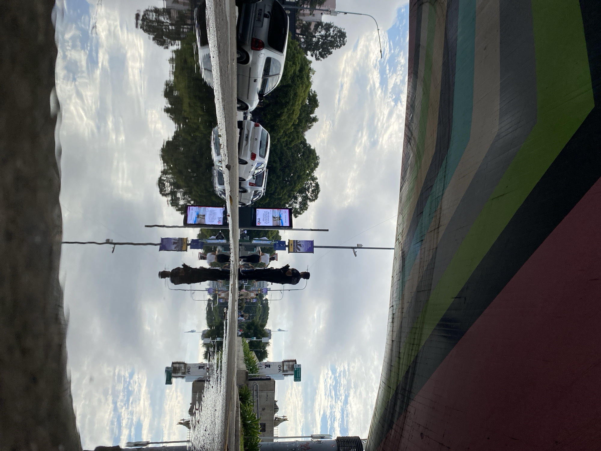 This image appears to be a reflection in a puddle on a street in Buenos Aires, Argentina. The upside-down reflection shows cars parked next to trees, street lights with banners, and people walking. On the right side of the frame, there is a portion of a bridge or wall that is painted with colorful stripes, including shades of green, gray, blue, and red. The sky is overcast.
