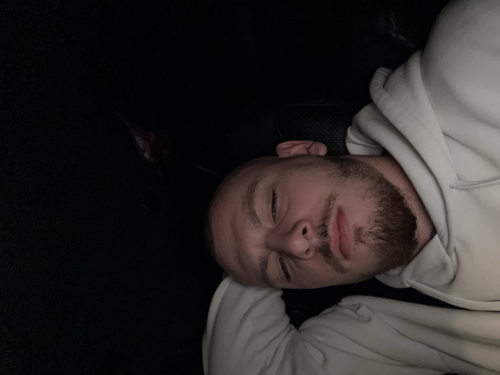 A man with a beard is lying on his side. He is wearing a white hoodie. The background is mostly dark, with what appears to be a car seat or other textured surface. The location of Stara Zagora, Bulgaria is not discernible in the image.