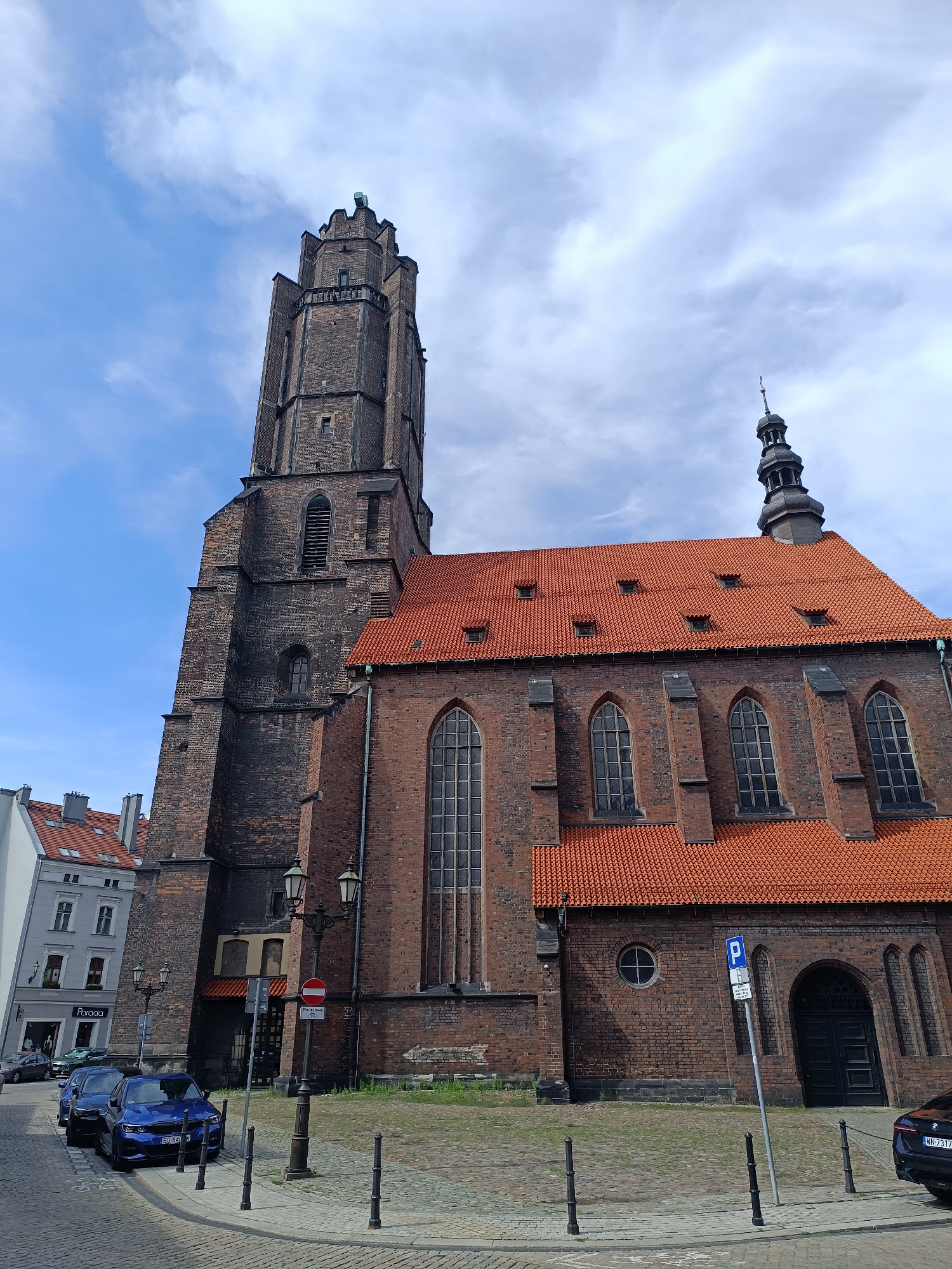 The image shows St. Bartholomew's Church (Kościół św. Bartłomieja) in Gliwice, Poland, a Gothic brick church with a tall, dark brick tower and a red-tiled roof. The church is situated on a cobblestone street, with several cars parked along the side. The sky is blue with scattered clouds. In the background, a few other buildings are visible, including a lighter-colored building with a shop on the ground floor. There are streetlights and bollards along the sidewalk. A parking sign is visible in front of the church.