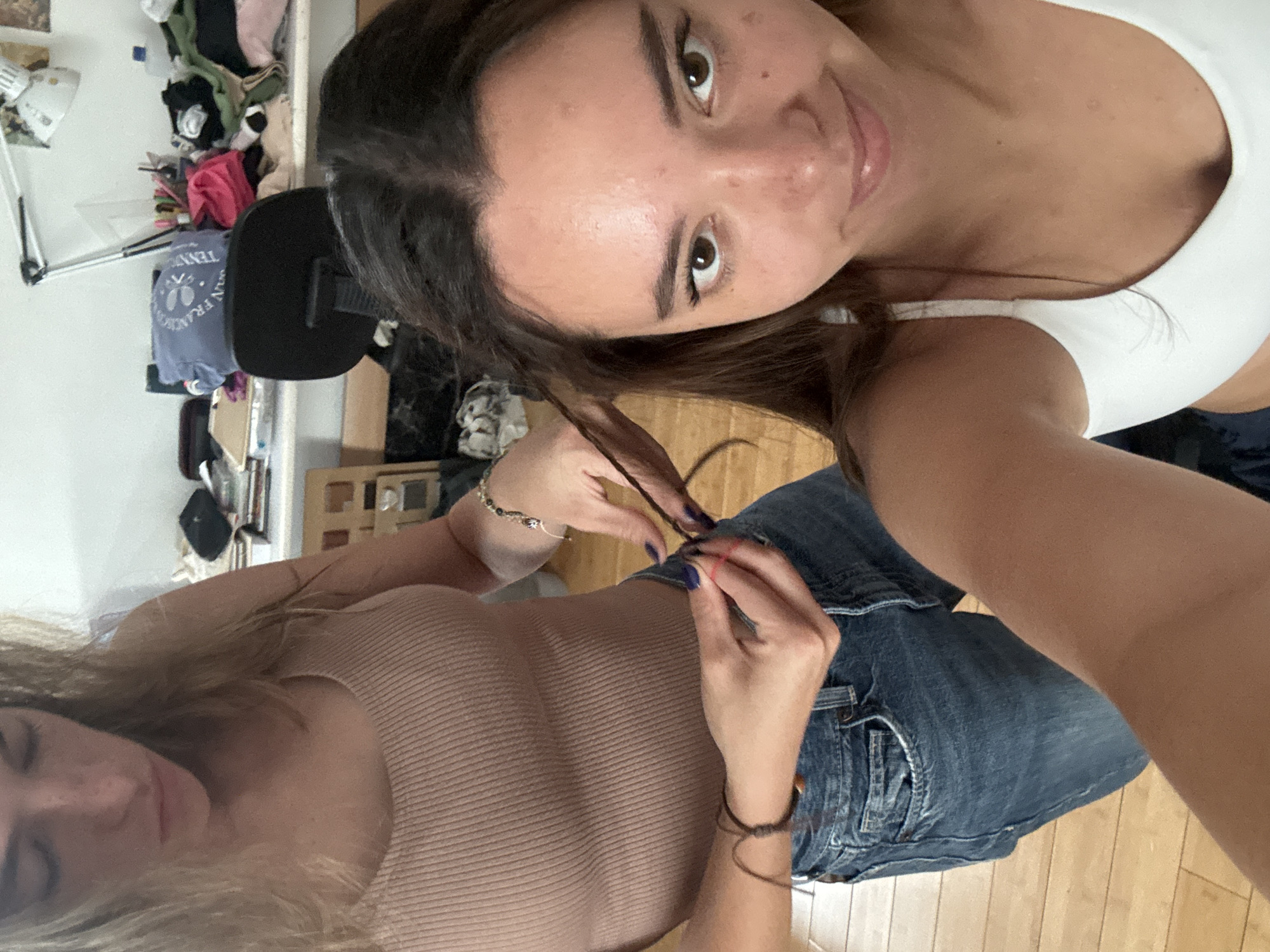 Two women are visible in an indoor setting. One woman is taking a selfie. She is wearing a white top. The second woman, wearing a beige top and blue jeans, appears to be braiding a strand of the first woman's brown hair. The location is potentially a home, evident by the presence of clothing and furniture in the background. The exact location in San Bernardino Tlaxcalancingo, Mexico cannot be determined from the image alone.