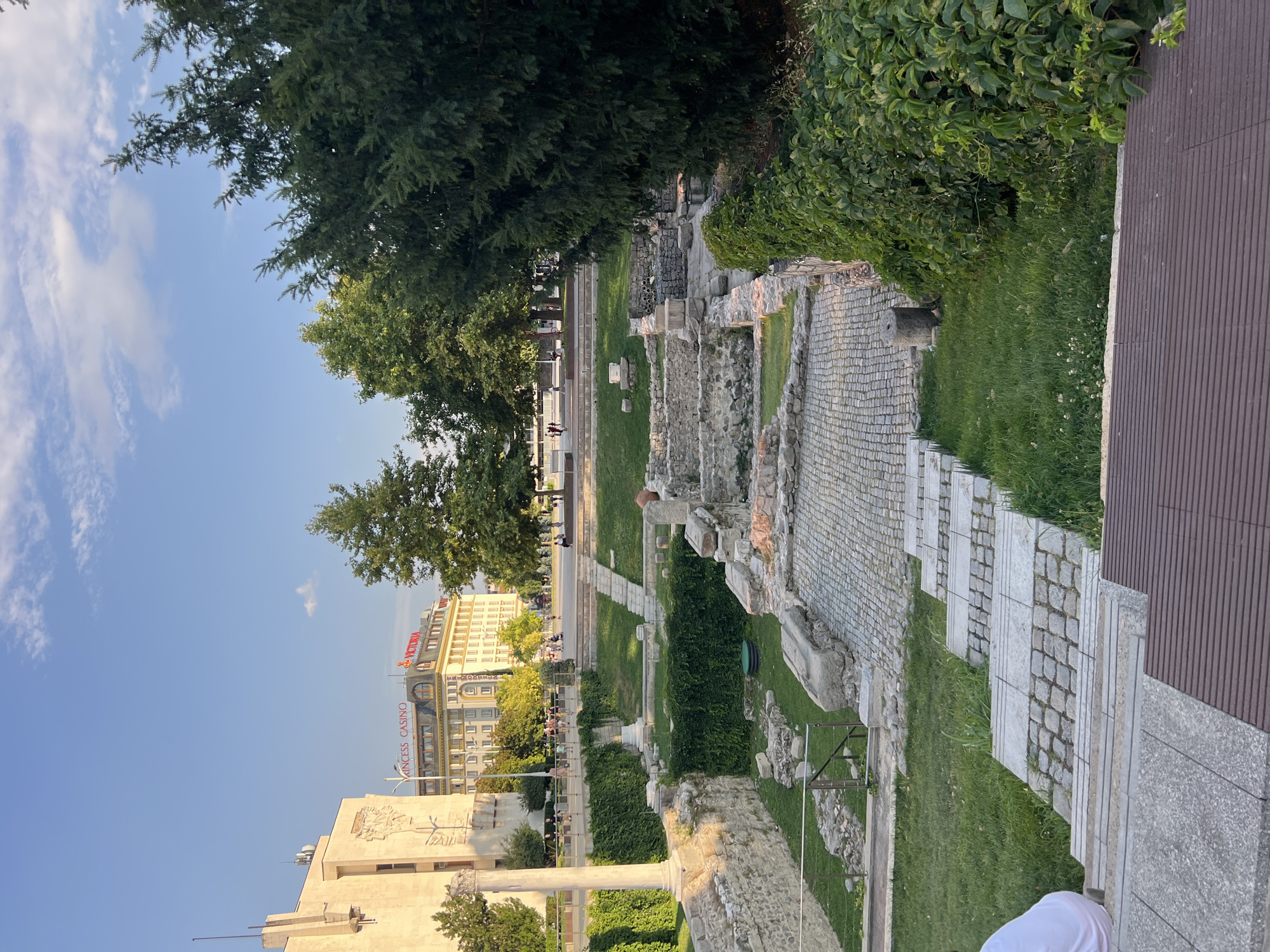 The image shows an archaeological site with ancient ruins in Plovdiv, Bulgaria, bathed in the warm light of a late afternoon sun. In the foreground, a person wearing a white shirt is seated on the edge of a wooden structure, looking down. A stone staircase, interspersed with cobblestone sections, leads into the excavated area.
The archaeological site features remnants of stone walls and structures, with cobblestone pathways winding through the ruins. Lush green grass covers the ground between the stone elements, and ivy vines climb along some of the walls. Beyond the ruins, a paved area with people walking and a line of trees separates the site from a street. In the background, a large, light-colored building with the words "PRINCESS CASINO" and "VICTORIA" in red letters is visible, along with other buildings and a clear blue sky with scattered clouds. The overall scene captures a blend of ancient history and modern city life.