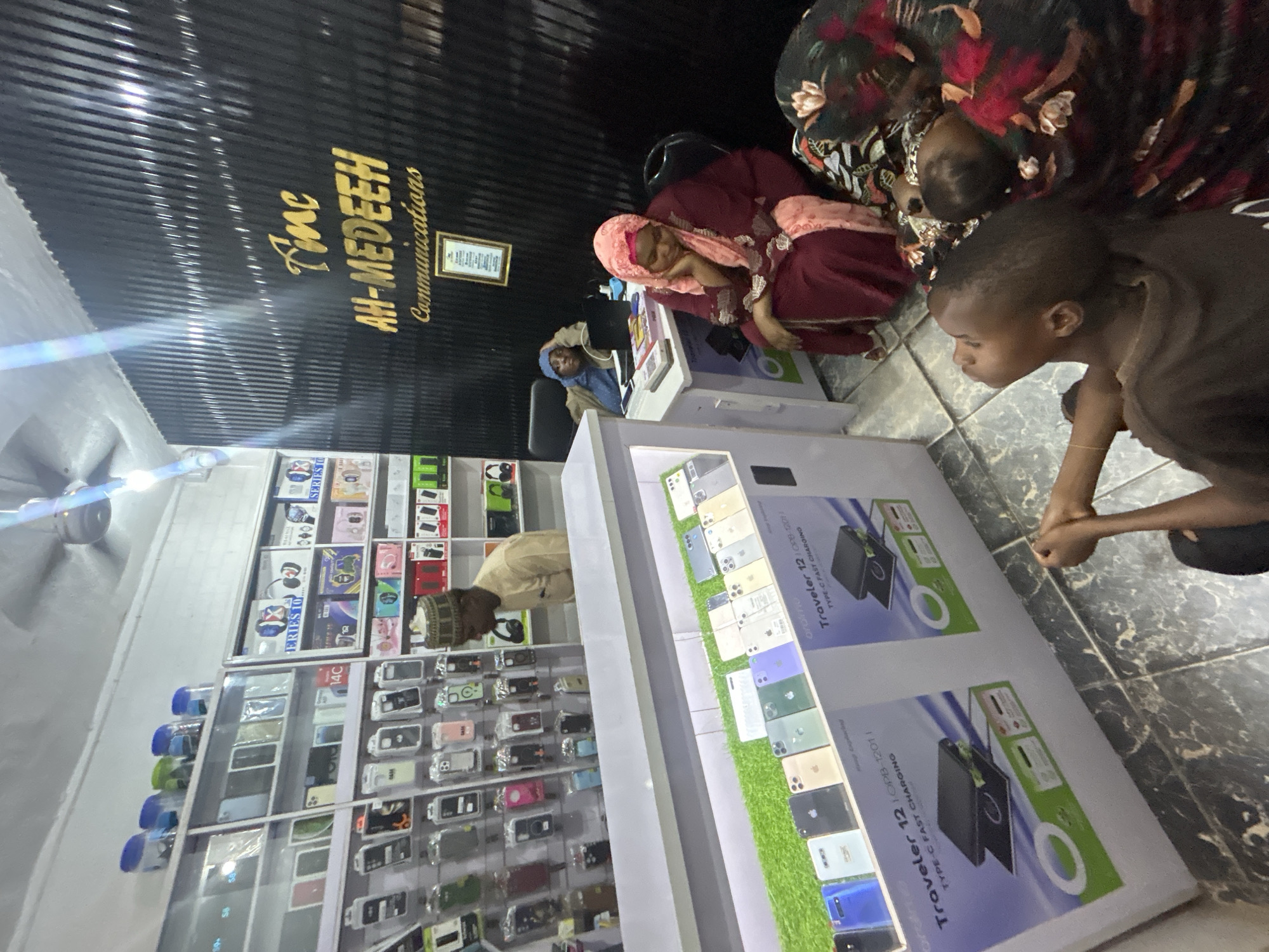 The interior of a small retail store called "AH-MEDEEH Communications" is depicted in a high-angle shot. A man wearing a white hat and a light-colored robe is examining items on a shelf loaded with phone cases and other accessories. Another person, possibly a shop assistant is seated behind a counter. A display case showcasing cellphones and charging accessories is in the foreground. A group of three people including a child is seated near the display case. The location is likely in Jos, Nigeria, as evidenced by the brand name of the store and the ethnicity of the people in the image.