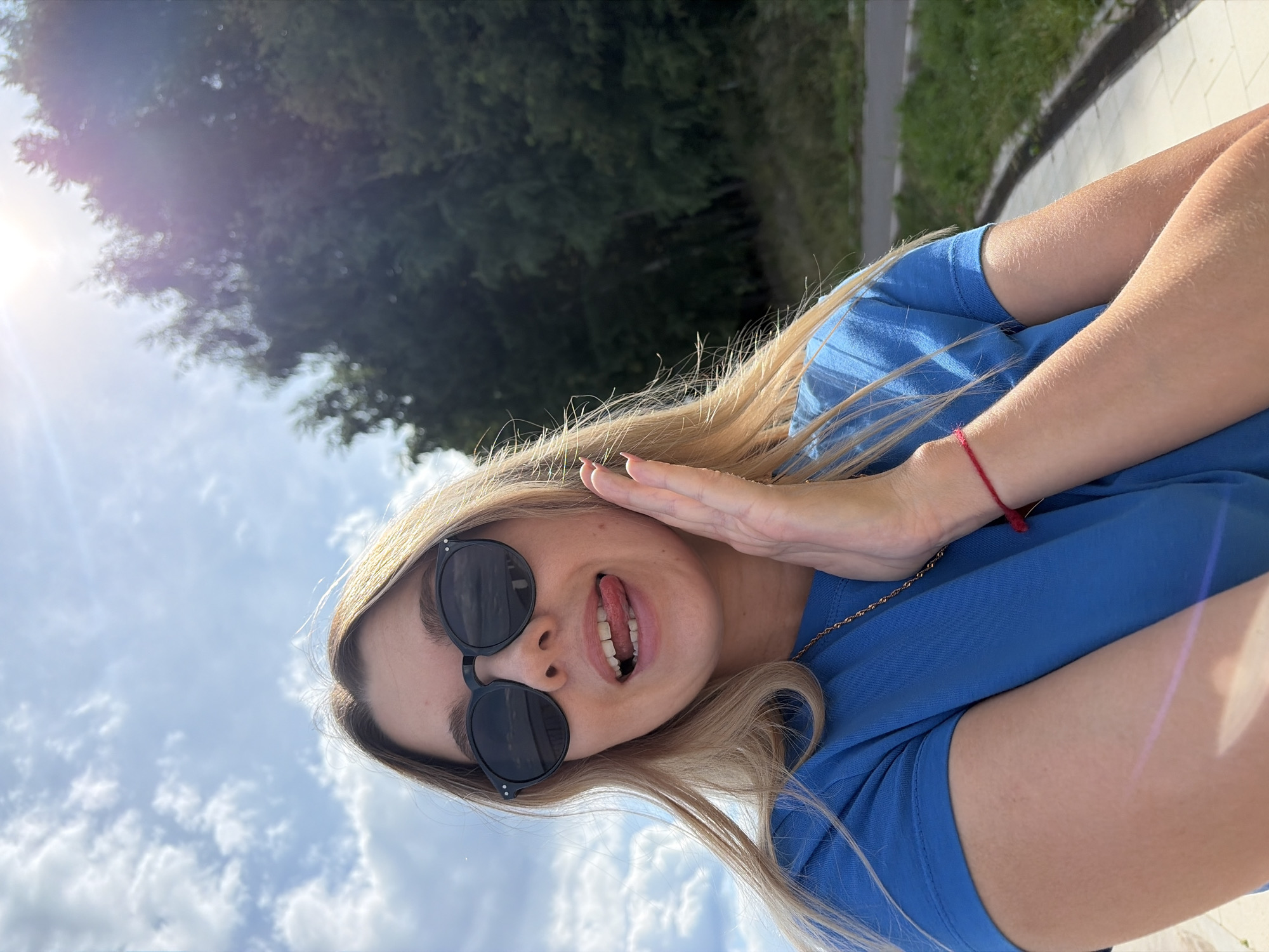 A young woman with blonde hair and sunglasses is taking a selfie outdoors on a sunny day. She is wearing a blue t-shirt and has her tongue sticking out. Her right hand is raised to her face. In the background, there are trees and a cloudy sky. The location is a park in Chaikovsky, Russia.