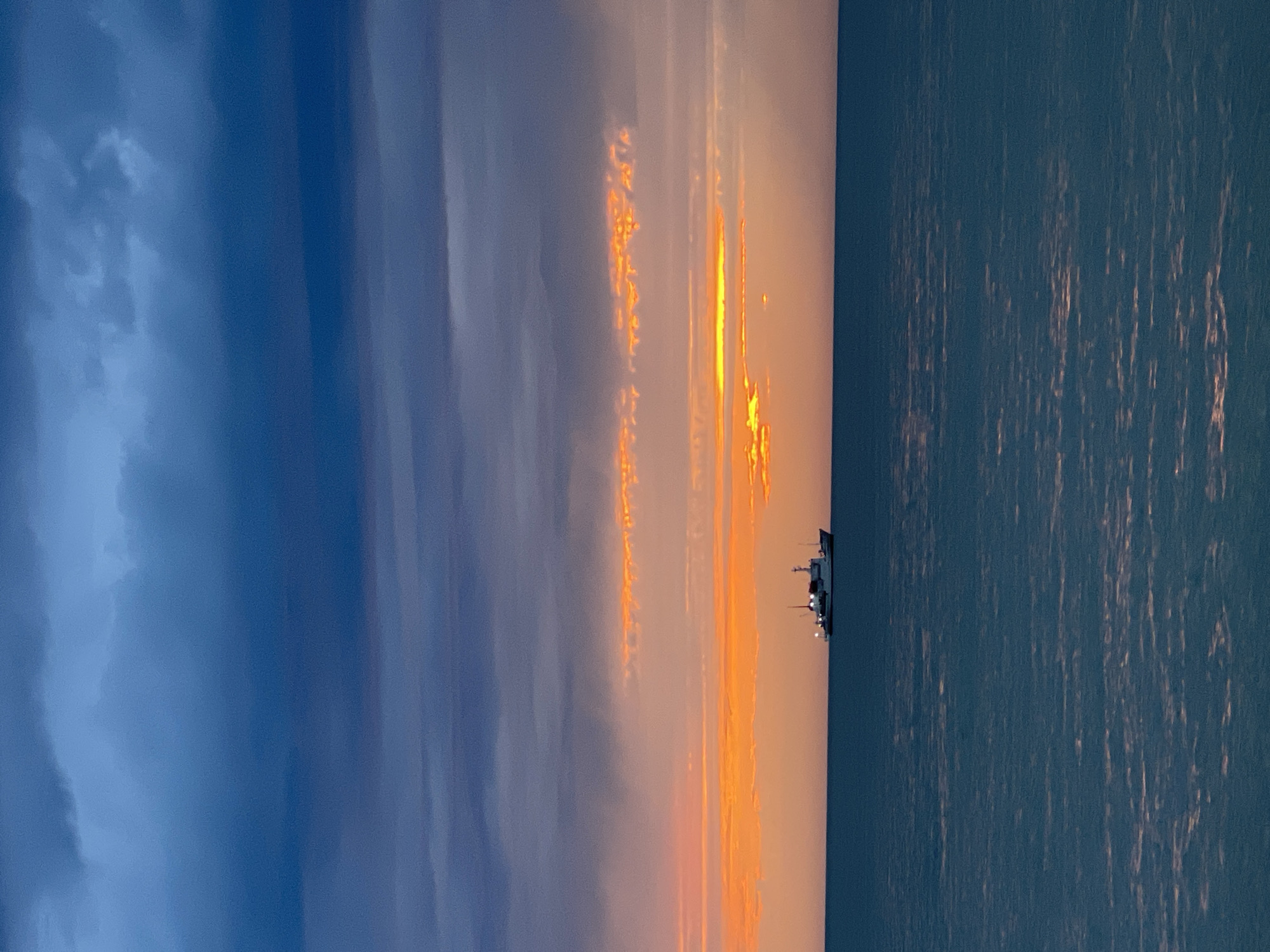 The image captures a sunset scene over the Black Sea, possibly near Sochi, Russia. The sky displays a gradient from dark blue to orange and peach hues where the sun is setting. Long vertical bands of orange-tinged clouds are present in the sky. A ship is visible on the horizon. The water reflects the light of the setting sun.