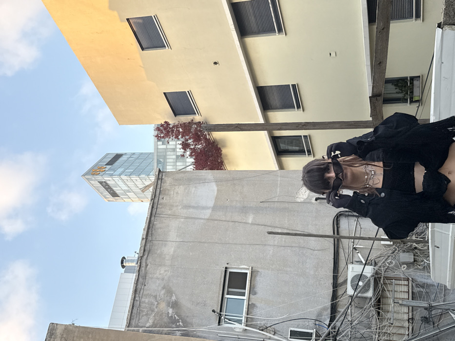 A young individual with dark hair and bangs stands in the foreground, wearing a black cropped top, a black fringed jacket, black gloves, and multiple layered necklaces. They hold dark rectangular sunglasses low on their nose, looking directly forward over the frames. The scene is an urban setting in Tel-Aviv, Israel. Behind the individual, a dilapidated, unpainted concrete building on the left exhibits a single horizontal window, exposed wires, and an external air conditioning unit. To the right, a multi-story, pale yellow building with dark external roller shutters on its windows is partially obscured by a vertical wooden beam. Between these older structures, a tree with red foliage is visible. In the background, the modern glass facade of the Azrieli Sarona Tower rises, identifiable by its distinctive orange hexagonal logo near the top. The sky above is partly cloudy blue.