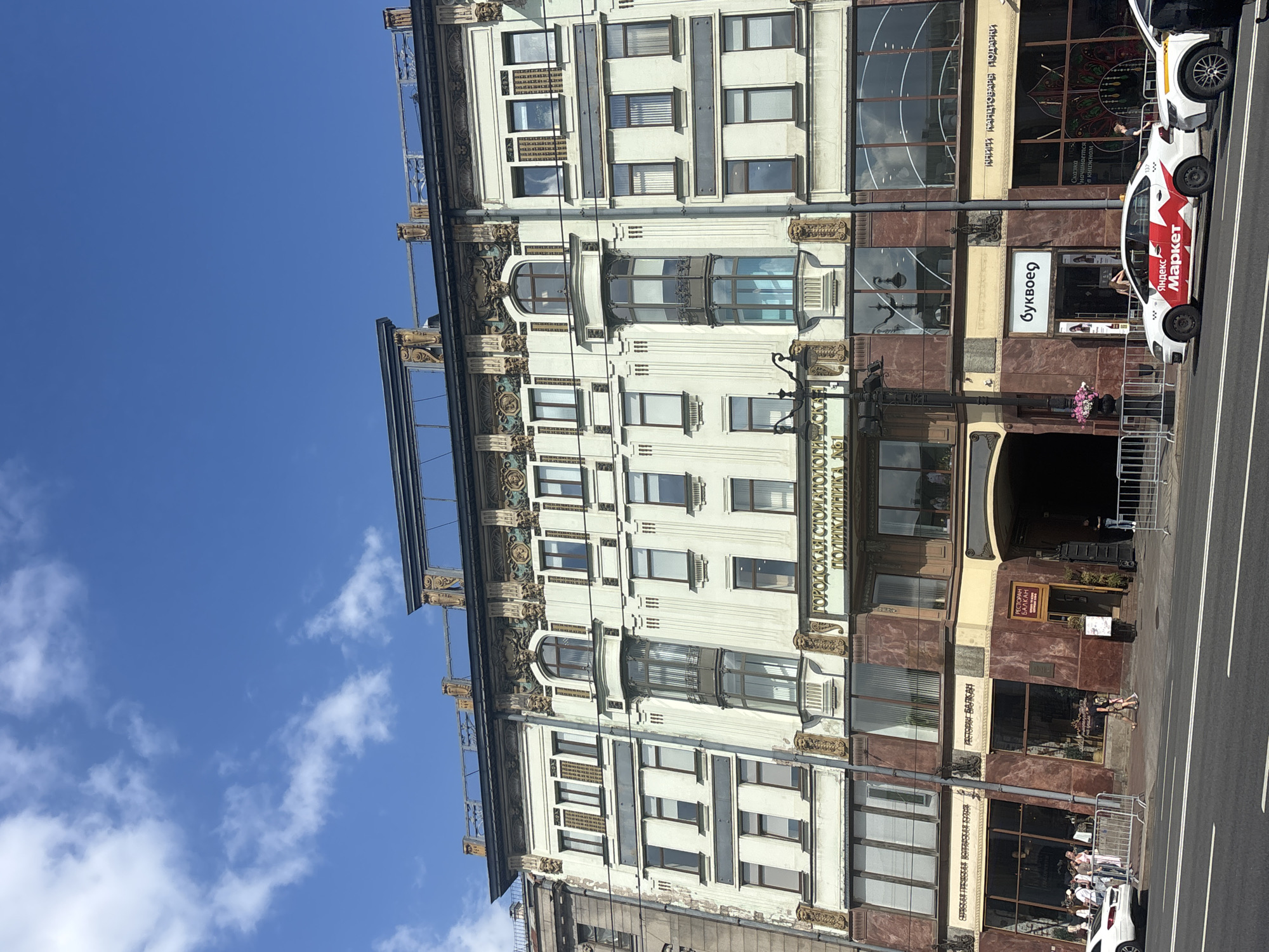 The image captures a street view of a building in Saint Petersburg, Russia, on a sunny day with a blue sky and scattered clouds. The building's facade is adorned with ornate architectural details, including decorative carvings and a multi-story design with numerous windows. Several businesses are visible at the ground level, including what appears to be a dental clinic ("ГОРОДСКАЯ СТОМАТОЛОГИЧЕСКАЯ ПОЛИКЛИНИКА №1"), a restaurant ("РЕСТОРАН БАЛКАН"), a bookstore ("Буквоед"), and a gift shop ("КНИГИ КАНЦТОВАРЫ ПОДАРКИ"). A white car with "Яндекс Маркет" branding is parked on the street in front of the building, along with other vehicles and pedestrians. The scene depicts a typical urban environment with commercial activity and public thoroughfares.