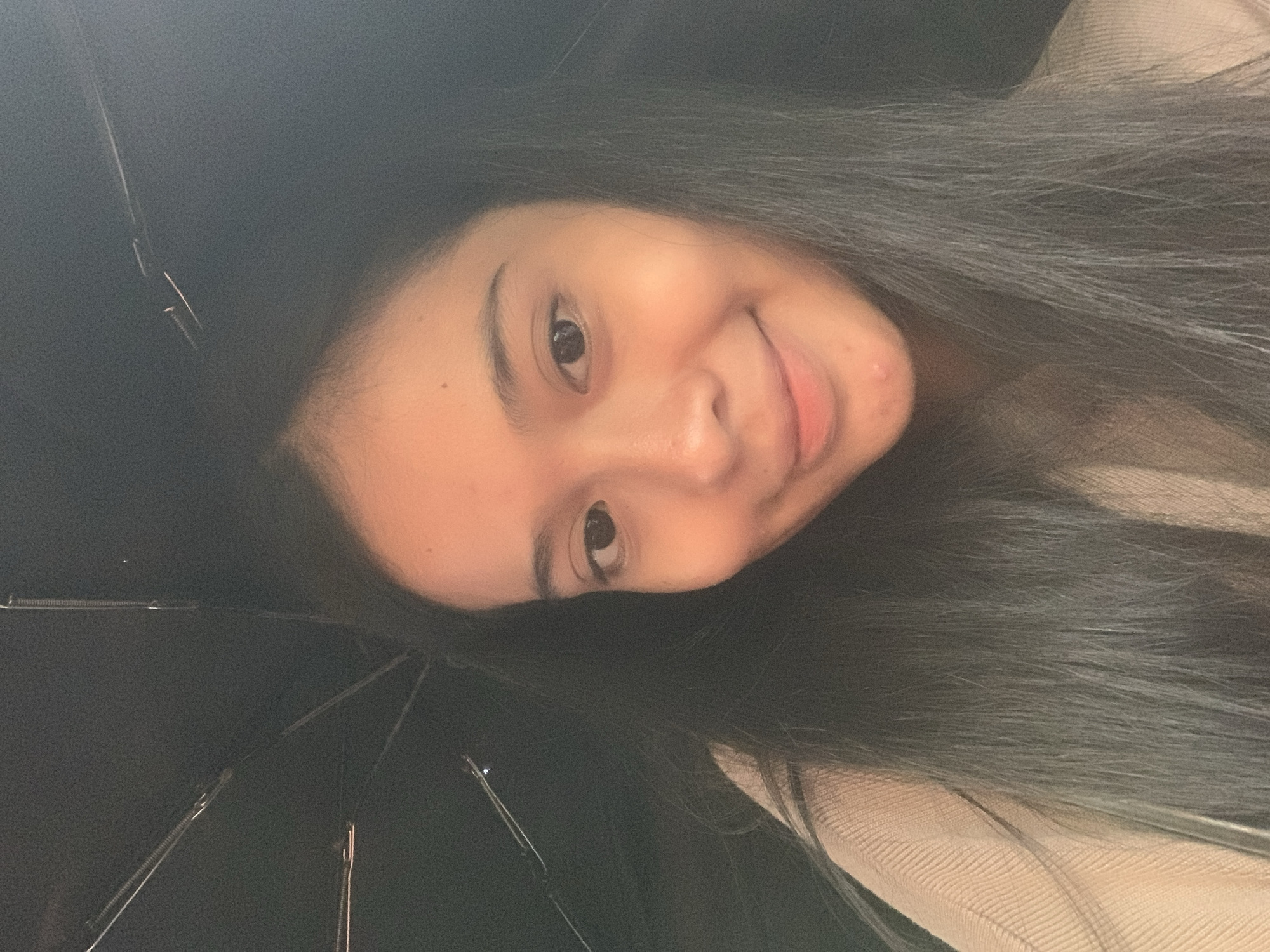 A young woman with long, dark hair is looking directly at the camera with a slight smile. She is partially obscured by a black umbrella, the metal spokes of which are visible in the lower and upper left corners of the frame. She is wearing a light beige sweater or top. The background is dark and indistinct. The location of Sungai Meriam, Indonesia is not apparent from the image.