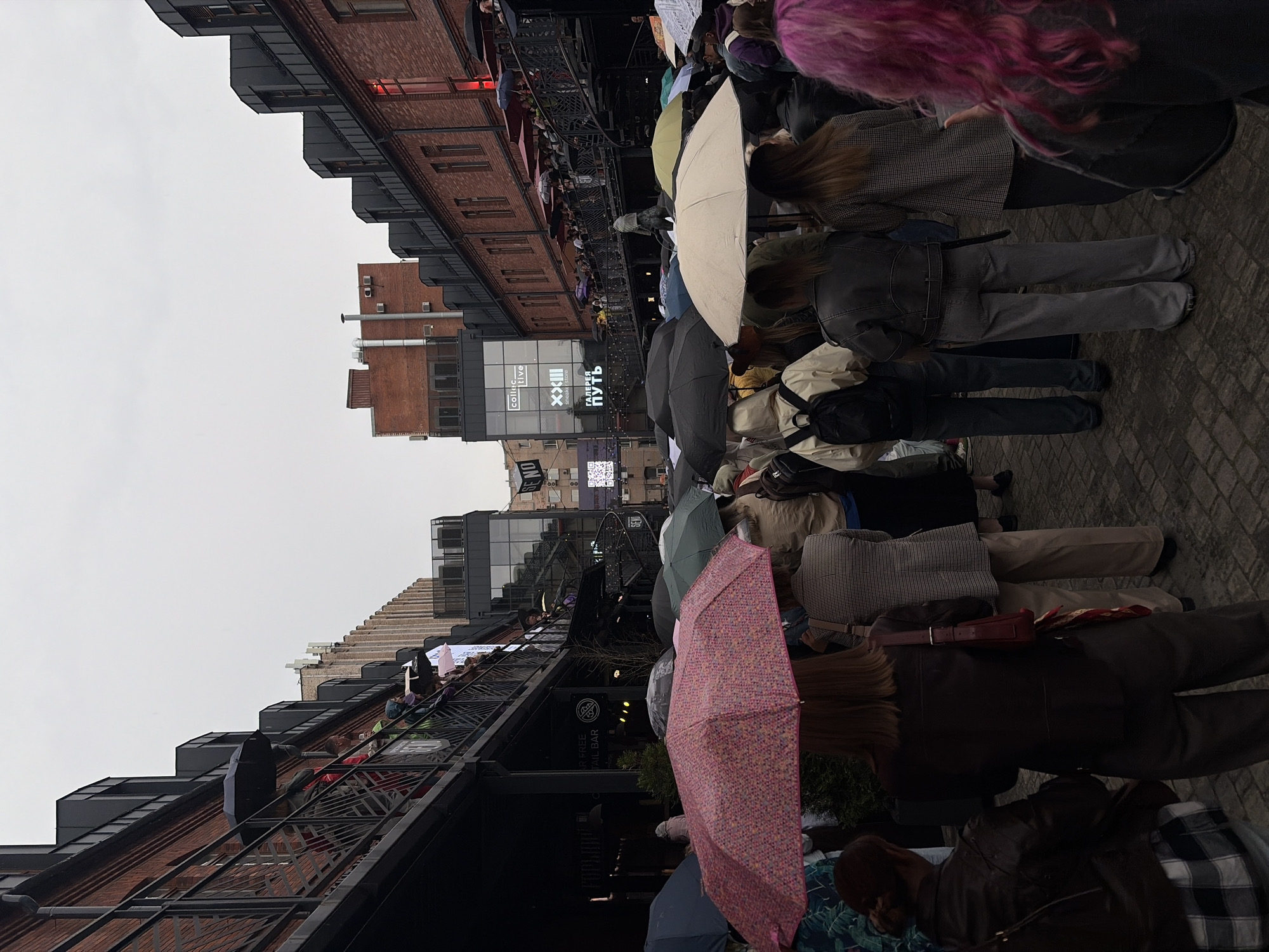 The image depicts a gathering of people under umbrellas on a cobblestone street in Saint Petersburg, Russia. The architecture suggests an urban environment, possibly a market or a district with industrial-chic buildings. Red brick buildings with black metal accents and balconies line the street. Many people are holding umbrellas, indicating it is raining. Some individuals are walking, while others appear to be standing and observing. There are signs on the buildings, one of which reads "XXIII SNEAKERSTORE." The overall atmosphere is muted due to the overcast sky and rain, with a focus on the people and their umbrellas.