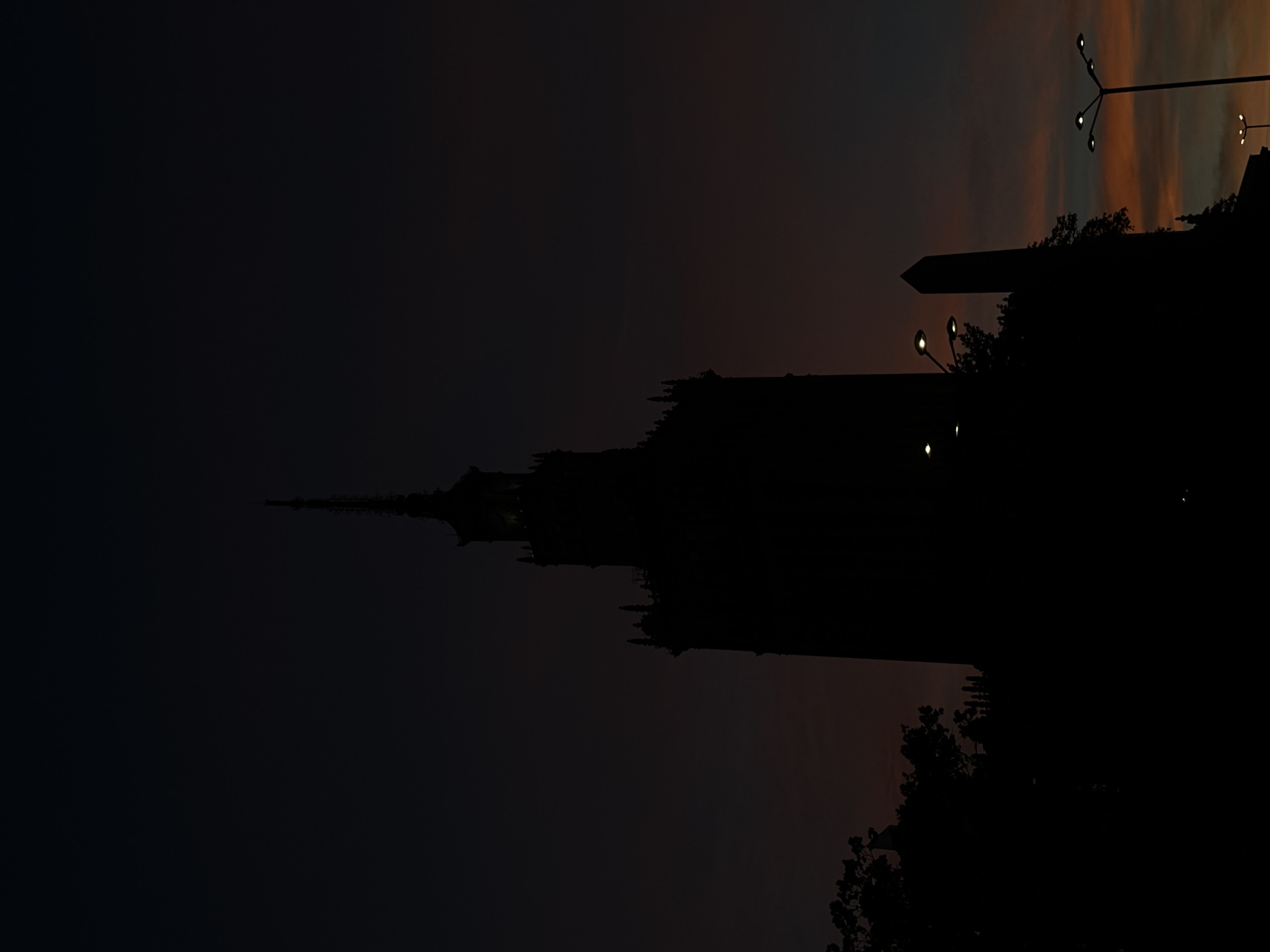The image is a silhouette of the Palace of Culture and Science in Warsaw, Poland, against a dark, twilight sky. The sky exhibits subtle hues of orange and dark blue, indicating the transition from sunset to night. The tall, gothic-style structure of the Palace dominates the central frame, its intricate spires and architectural details rendered in black. To the left of the Palace, the silhouette of trees is visible, adding a natural element to the urban landscape. Several streetlights are scattered throughout the scene, with their bright bulbs providing small points of illumination against the dark surroundings. One streetlight, in particular, is prominently positioned in the lower right quadrant of the image. A slender, obelisk-like monument stands to the right of the Palace, also in silhouette. The overall mood is somber and dramatic, emphasizing the imposing presence of the iconic building at dusk.