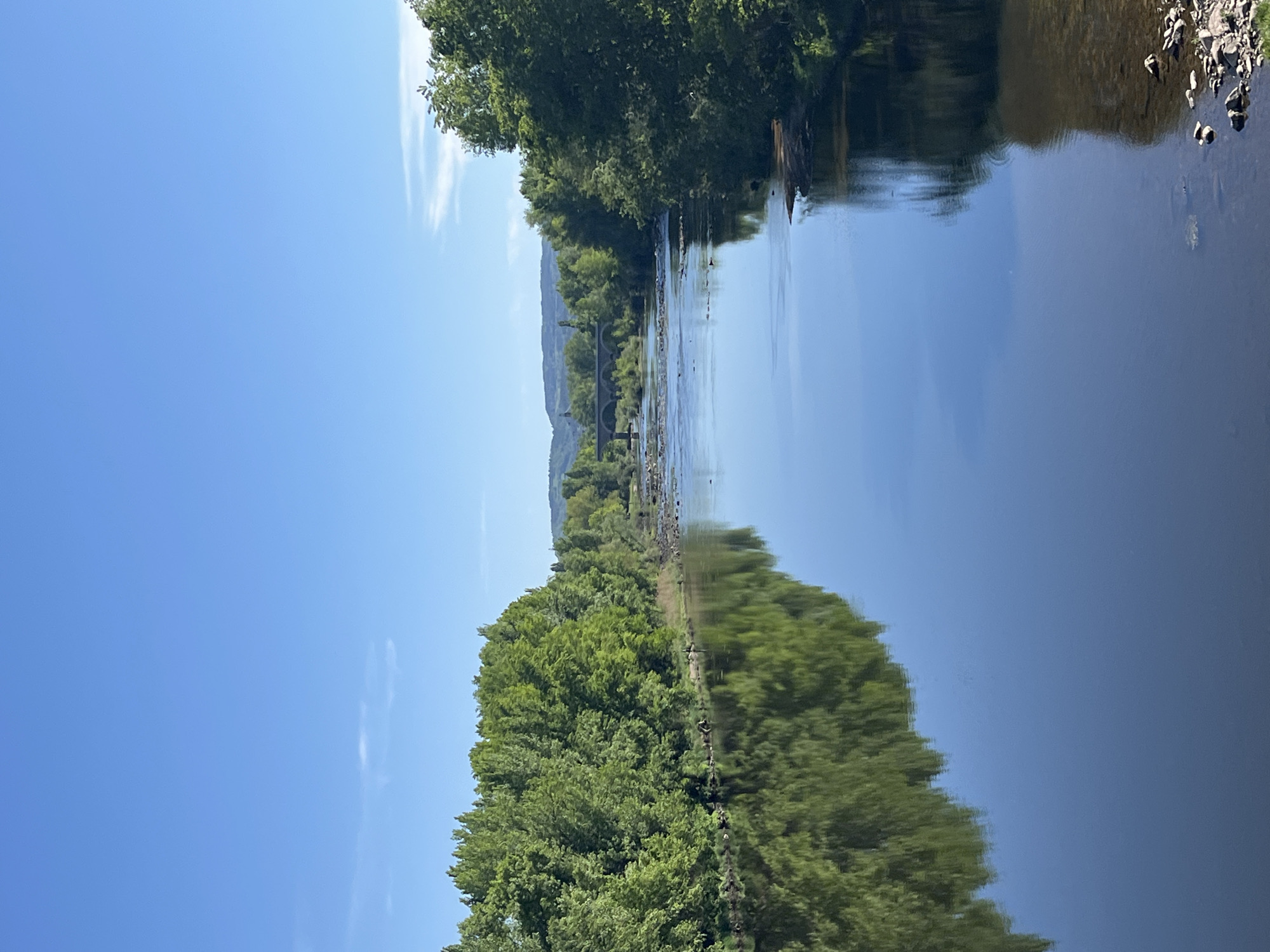 The image depicts a river scene in Andrézieux-Bouthéon, France. The river reflects the sky and the lush green trees lining its banks, creating a mirrored effect. A bridge is visible in the distance. The sky is clear and blue with a few wispy clouds. The right bank has a rocky area where land meets the water.
