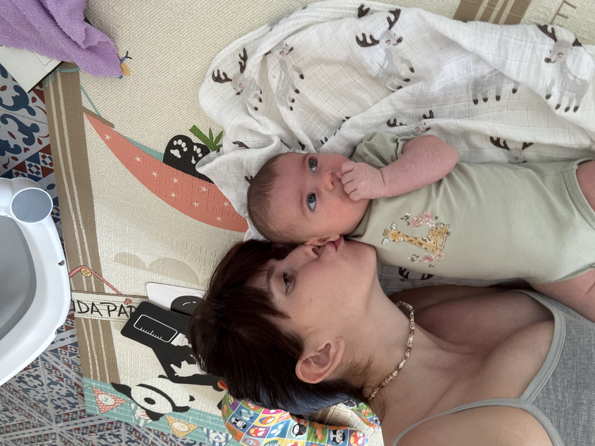 A woman and a baby are lying on a play mat. The woman is kissing the baby on the cheek. The baby is wearing a light green onesie with a giraffe print. The woman is wearing a grey tank top and a beaded necklace. There is a purple cloth and a white blanket with reindeer prints on the play mat. The play mat has various colorful patterns and cartoon characters, including pandas and a hammock with paw prints. In the background, a white object with a blue button is visible, possibly a baby monitor or bath toy. The scene appears to be indoors, likely a playroom or nursery. The lighting suggests daytime. The overall mood is affectionate and tender, conveying a sense of love and closeness between mother and child. Text visible on the play mat reads "DA PAT."