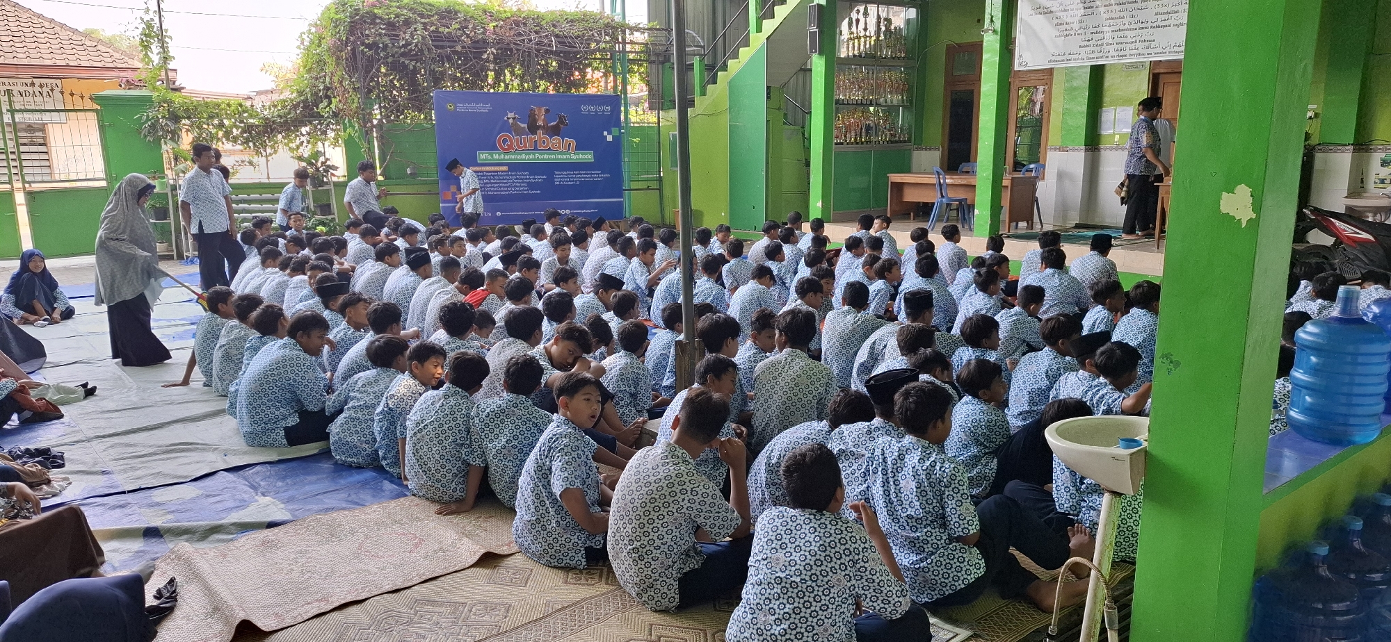 In Wonorejo, Indonesia, a large group of young boys, most wearing matching blue and white patterned shirts and black pants or sarongs, are seated on mats on the ground. They appear to be students at an Islamic school, possibly during an outdoor assembly or class. A woman in a gray headscarf and black robe stands to the left, holding a broom or stick. In the background, a blue banner with Arabic text and images of cows is displayed, along with a green building and stairs. Some students are looking towards the front, while others are interacting with each other. The environment suggests a warm, sunny day.