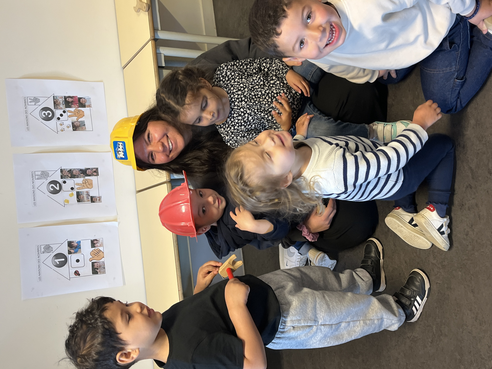 A medium shot shows a group of adults and children. A woman with dark hair wears a yellow hard hat and smiles. A child in a red hard hat holds a small wooden hammer toy. The others wear normal clothes. Three sheets of paper are affixed to the wall. The sheets feature a house with a number on it and images of dice, fingers, and faces inside. The number of the house progresses from 1 to 3. Text above each house says, "LES MAISONS DES NOMBRES". The image was taken in La Fouillouse, France.