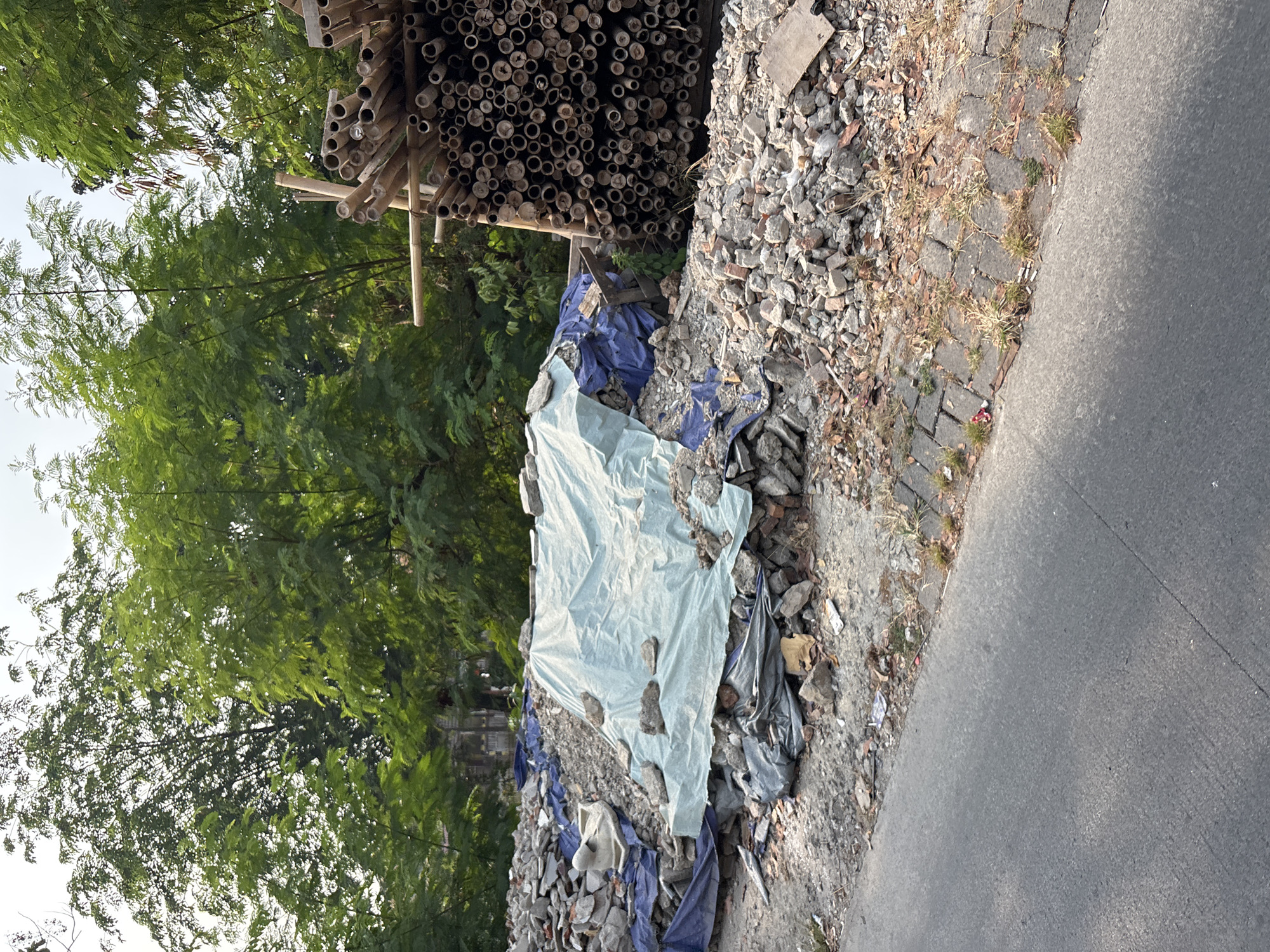 A pile of construction debris, including broken concrete, bricks, and a partial white sink, is covered in blue and light blue tarps along a paved road. To the right of the debris is a large stack of bamboo poles of varying diameters, bound together. Behind the debris and bamboo, dense green foliage from trees creates a backdrop. The scene appears to be in a semi-urban or developing area, consistent with locations in Bekasi, Indonesia, where such construction and material staging are common. The lighting suggests it is daytime, possibly late afternoon, given the shadows. No people are visible in the frame.