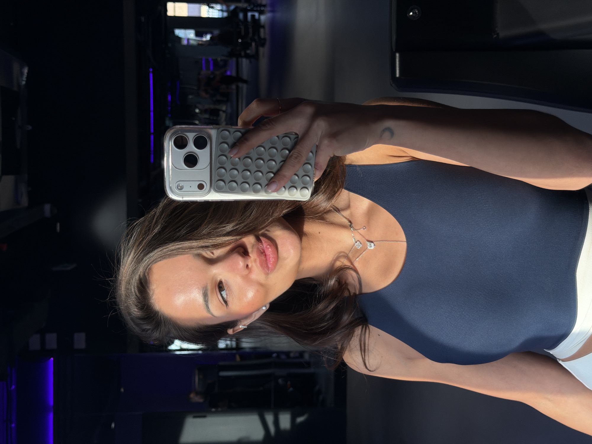 This image captures a mirror selfie of a woman in what appears to be a gym or fitness center in Krasnoyarsk, Russia. The woman is positioned with her head slightly tilted and a subtle, confident smile. She has long, dark brown hair and is wearing a dark blue athletic top, with a glimpse of white athletic bottoms visible.
She is adorned with delicate jewelry, including two necklaces—one featuring a clover-like design with clear stones, and another with a small, sparkling pendant—as well as small stud earrings. A small crescent moon tattoo is visible on the inner side of her right forearm.
The woman is holding a smartphone in her right hand, facing her reflection to capture the selfie. The phone is encased in a distinctive light-colored, transparent case with a grey, textured back panel composed of numerous small, raised circular bumps, likely for enhanced grip.
The background is dark and blurred, revealing typical gym equipment and subtle purple LED lighting, suggesting an indoor setting. Strong, directional lighting illuminates the woman's face and right shoulder, creating highlights and shadows. No discernible text is visible within the image.