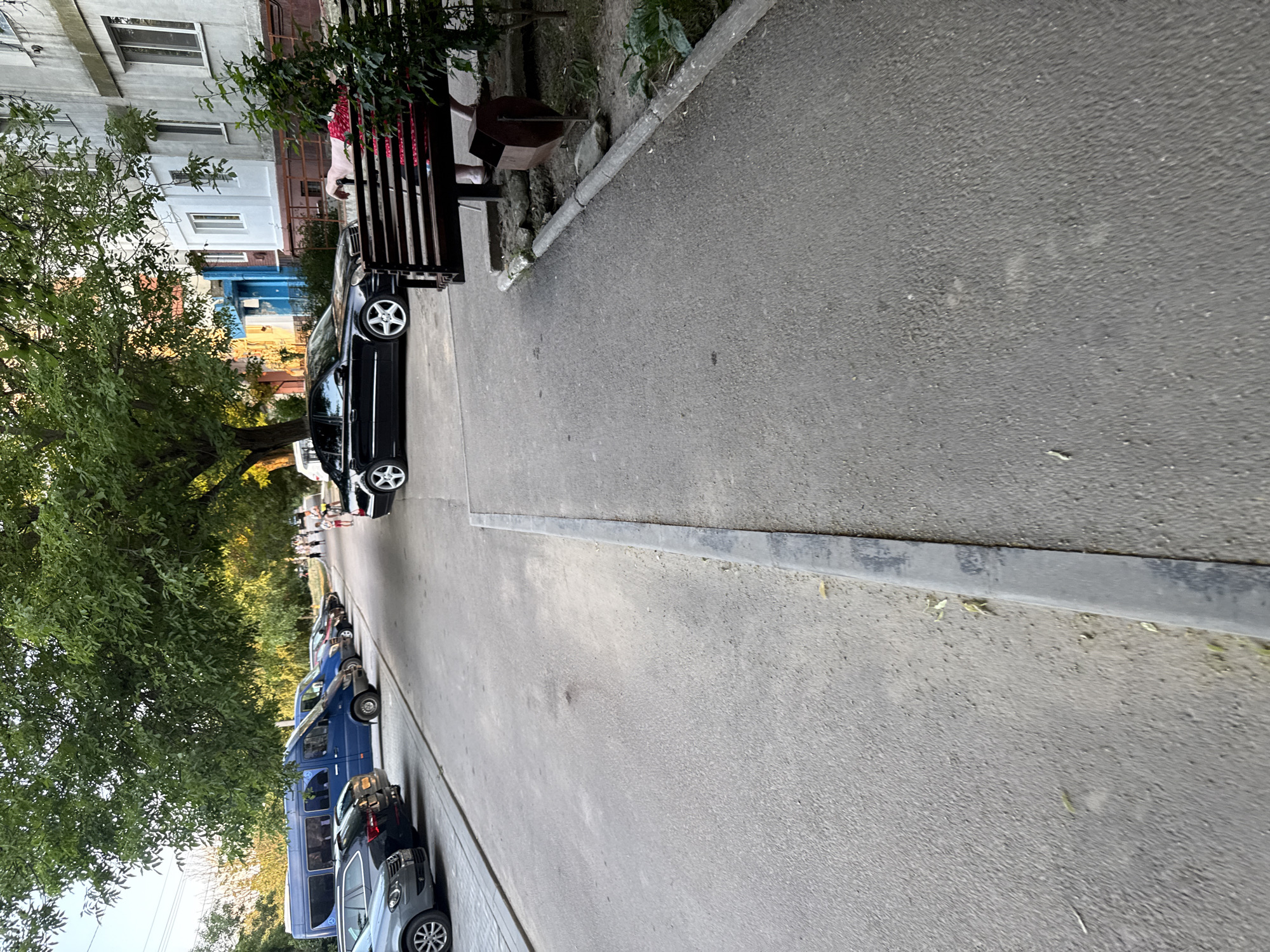 A street scene in Cahul, Moldova, shows parked cars, a blue minibus, and a black sedan. A tree with dense green foliage provides shade over the street. The street is paved and has a raised sidewalk along one side. A wooden bench is situated on the sidewalk, and a person wearing a red dress is seated on it. A metal trash receptacle is located next to the bench. Buildings with apartment windows are visible in the background.