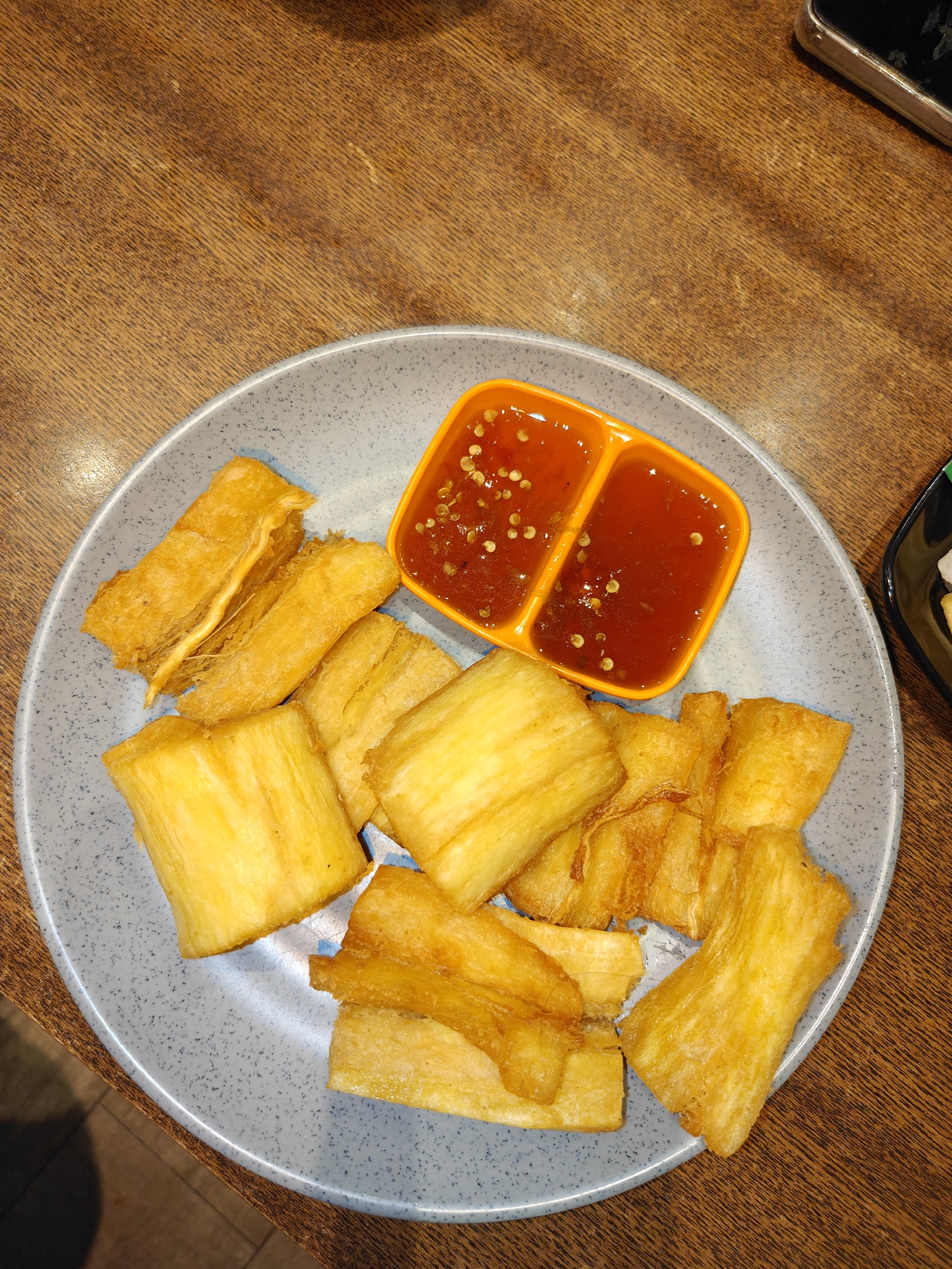 A plate of fried cassava sticks and dipping sauce sits on a wooden table. The cassava is golden brown and appears crispy. The dipping sauce is red and appears to be sweet chili sauce. There is also a sliver of black dish with some white food on it visible on the right side of the frame. The table is made of wood and has a visible grain pattern. The lighting is warm and appears to be natural or from overhead lights. No people are visible in the image. The scene is a close-up, top-down view of the food and table setting. The location of Situbondo, Indonesia is inferred from context not visible in the image itself.
