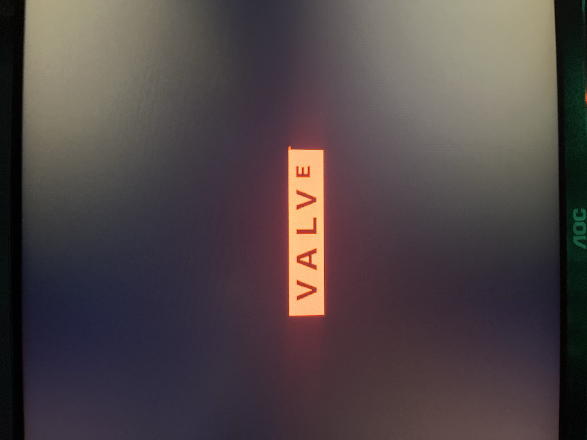 The image shows a computer screen displaying the word "VALVE" in red text on an orange rectangular background. The background of the screen is a gradient of dark blue to light gray, with the bottom edge of the screen showing the "AOC" logo in green. There is no indication of the location being Chaikovsky, Russia, nor are there any people or actions depicted.