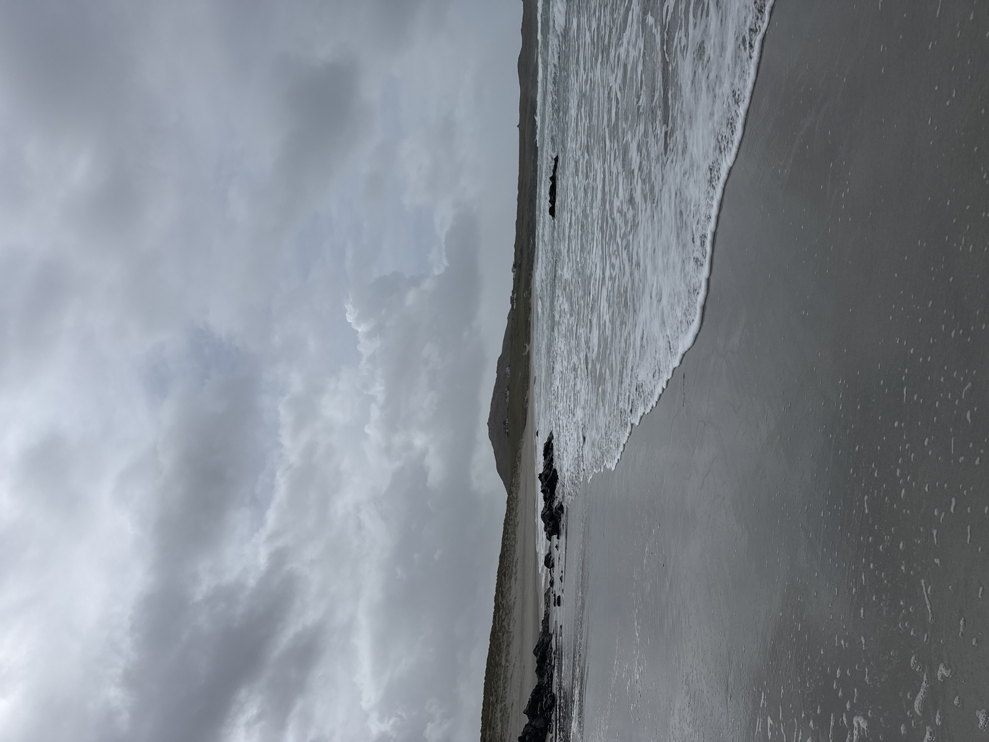 The image shows a grayscale beach scene, likely taken on an overcast day. The sky is filled with dense, gray clouds. The beach slopes down towards the ocean, with the waves washing onto the shore. Dark rocks are scattered along the shoreline. A hill or mountain is visible in the distance, partially obscured by the clouds. The location is unknown.