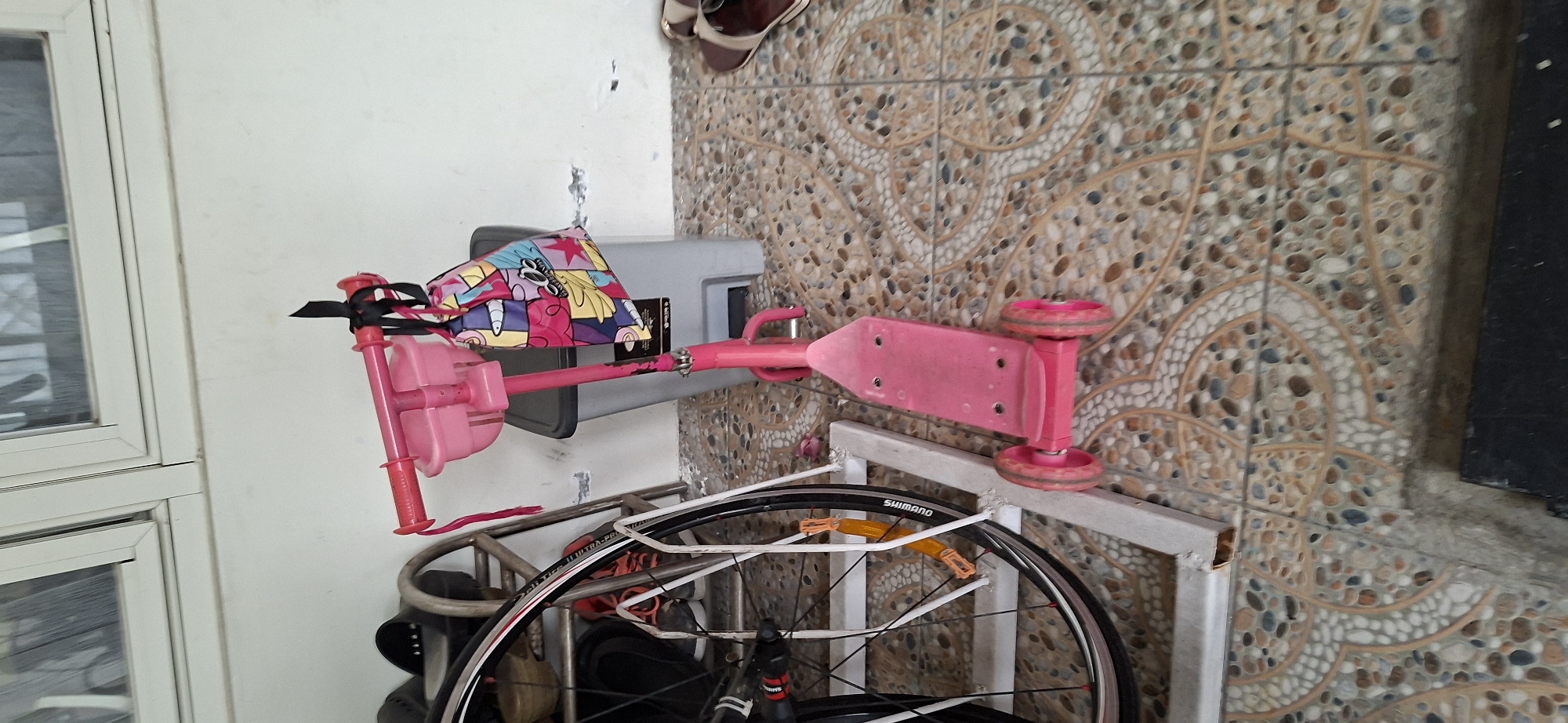 A pink three-wheeled scooter with a patterned bag attached to the handlebars stands on a tiled floor. To the left of the scooter, a bicycle wheel is propped up against a metal stand. In the background, a white wall and a window with horizontal blinds are visible. The scene appears to be an outdoor covered area, possibly a porch or carport, based on the presence of the tiled floor and the wall. The image is taken from a slightly high angle, looking down at the scooter. The lighting suggests it is daytime. This image is located in Surabaya, Indonesia.
