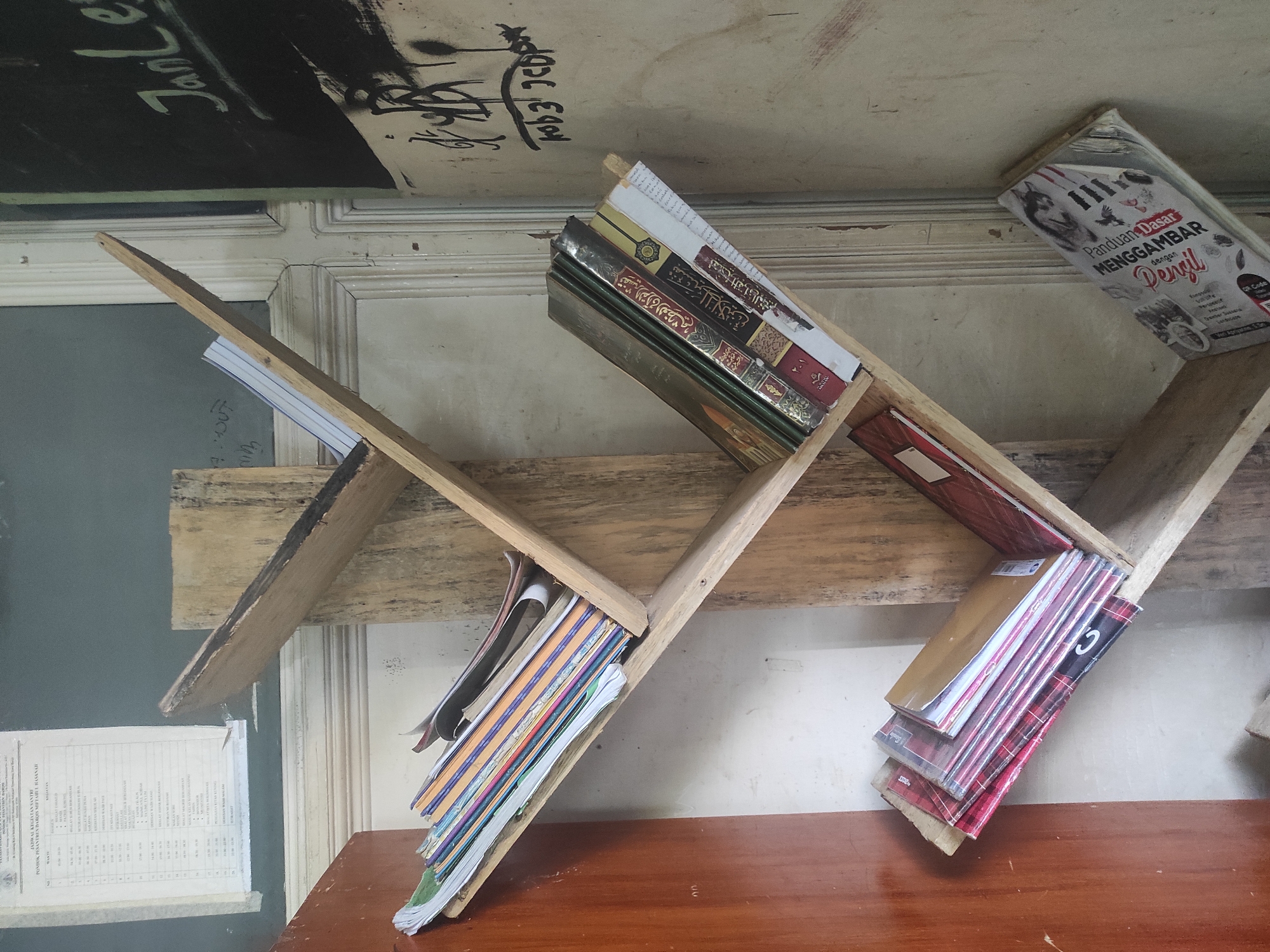 The image depicts an interior scene, likely a classroom or study area, featuring a rustic, DIY wooden bookshelf. The bookshelf is made from rough-hewn planks and is positioned against a wall with a patterned texture. Various books and notebooks are stored on the shelves.
On the uppermost shelf, a stack of thin notebooks or papers is visible. The second shelf from the top holds several books, some with colorful spines. The third shelf contains a row of books with Arabic script on their covers, suggesting religious texts. The fourth shelf down displays a stack of notebooks with plaid covers. The bottom shelf has a single, open book with a title that appears to be in Indonesian.
To the left of the bookshelf, a section of a wooden door or cabinet is visible. In the background, a blackboard or dark-colored wall is partially visible, with some writing in white chalk. The overall environment appears somewhat aged and worn.
While the image does not explicitly indicate the city of Sumedang, Indonesia, the presence of Indonesian text on one of the books suggests a possible location within Indonesia. The scene evokes a sense of a functional, albeit unpolished, learning or communal space.