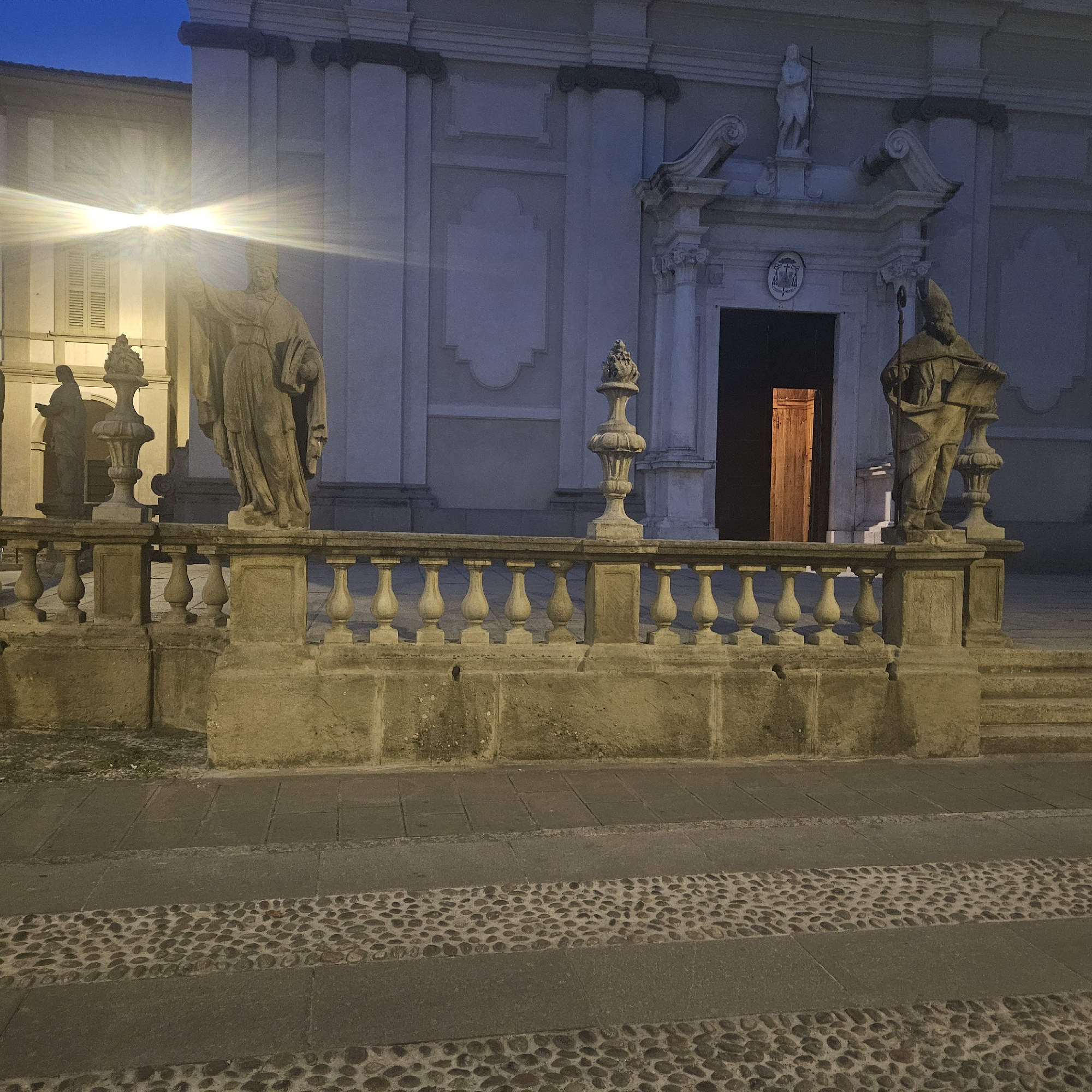 The image shows the facade of the church of San Giovanni Battista in Stezzano, Italy at dusk. The scene includes a stone balustrade with statues of religious figures on either side of the entrance. A light source shines brightly, illuminating the statue on the left side. The entrance to the church is visible, with an open door revealing a warmly lit interior. The ground is paved with a pattern of stone and cobblestone. The building's architecture is characterized by classical elements.