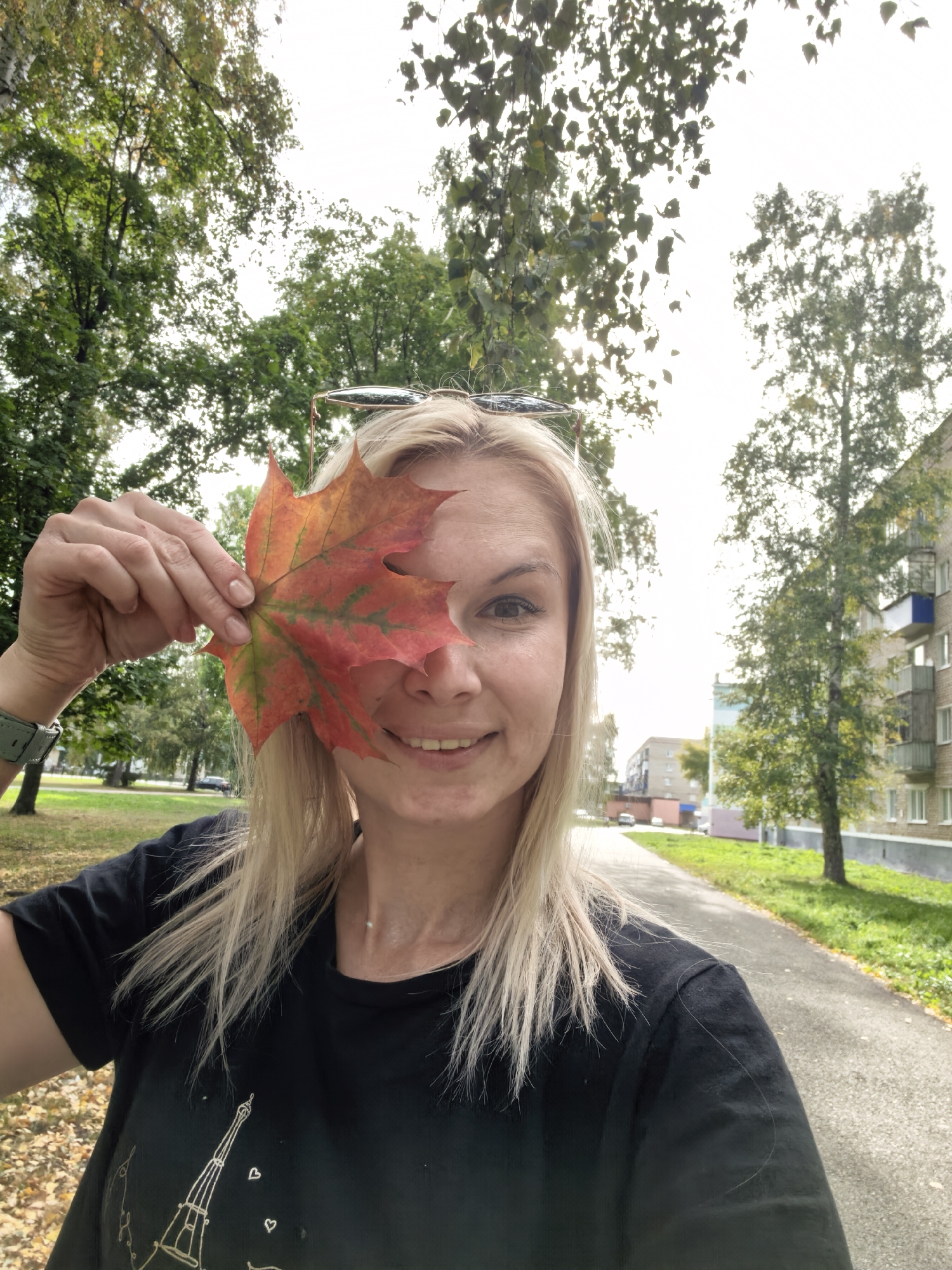 In Sterlitamak, Russia, a blonde woman with fair skin is taking a selfie outdoors. She is holding a vibrant red and yellow maple leaf in front of her face, obscuring her left eye and part of her cheek. She is smiling, revealing her teeth, and her right eye is visible, looking towards the camera. She has blonde hair, and sunglasses are perched on top of her head. She is wearing a black t-shirt with a white graphic of the Eiffel Tower and small hearts. A thin necklace with a small pendant is visible around her neck. She is also wearing a watch on her left wrist. The background features a paved walkway, green grass, trees with green and autumn-colored leaves, and a brick apartment building with balconies. The sky is overcast, with bright sunlight filtering through the clouds. The leaves on the ground suggest it is autumn.