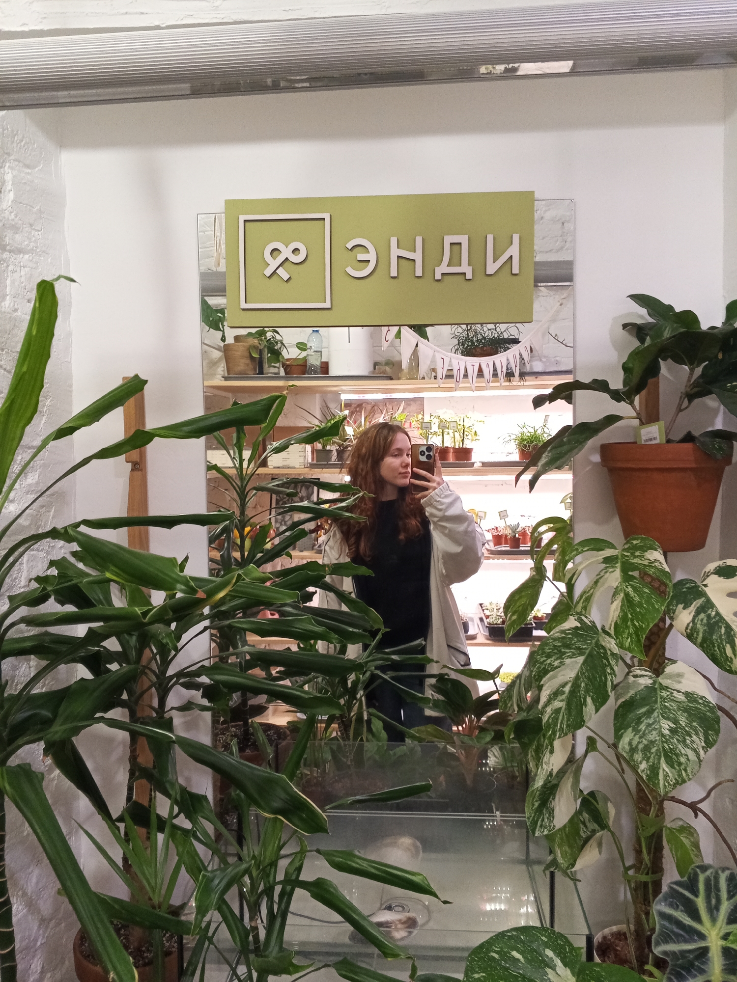 The image depicts an indoor scene within a plant store, specifically at ENDI Houseplant and Terrarium Store located at Apraksin Dvor, Sadovaya Ulitsa 28-30, Saint Petersburg, Russia. A young female individual with long, wavy reddish-brown hair is captured in a mirror reflection, taking a selfie. She is wearing a light-colored jacket over a dark top and holds a dark-colored smartphone with multiple rear cameras. The mirror occupies a significant portion of the frame, reflecting shelves laden with various potted plants, some illuminated by grow lights. Above the reflected shelving, a light-green sign displays a white infinity symbol-like logo within a square frame, alongside the Cyrillic text "ЭНДИ" (ENDI). The foreground and left side of the image are filled with live plants, including large green specimens and a prominent variegated Monstera plant with white and green leaves. The immediate environment features white walls, one exhibiting a textured, possibly painted brick surface.