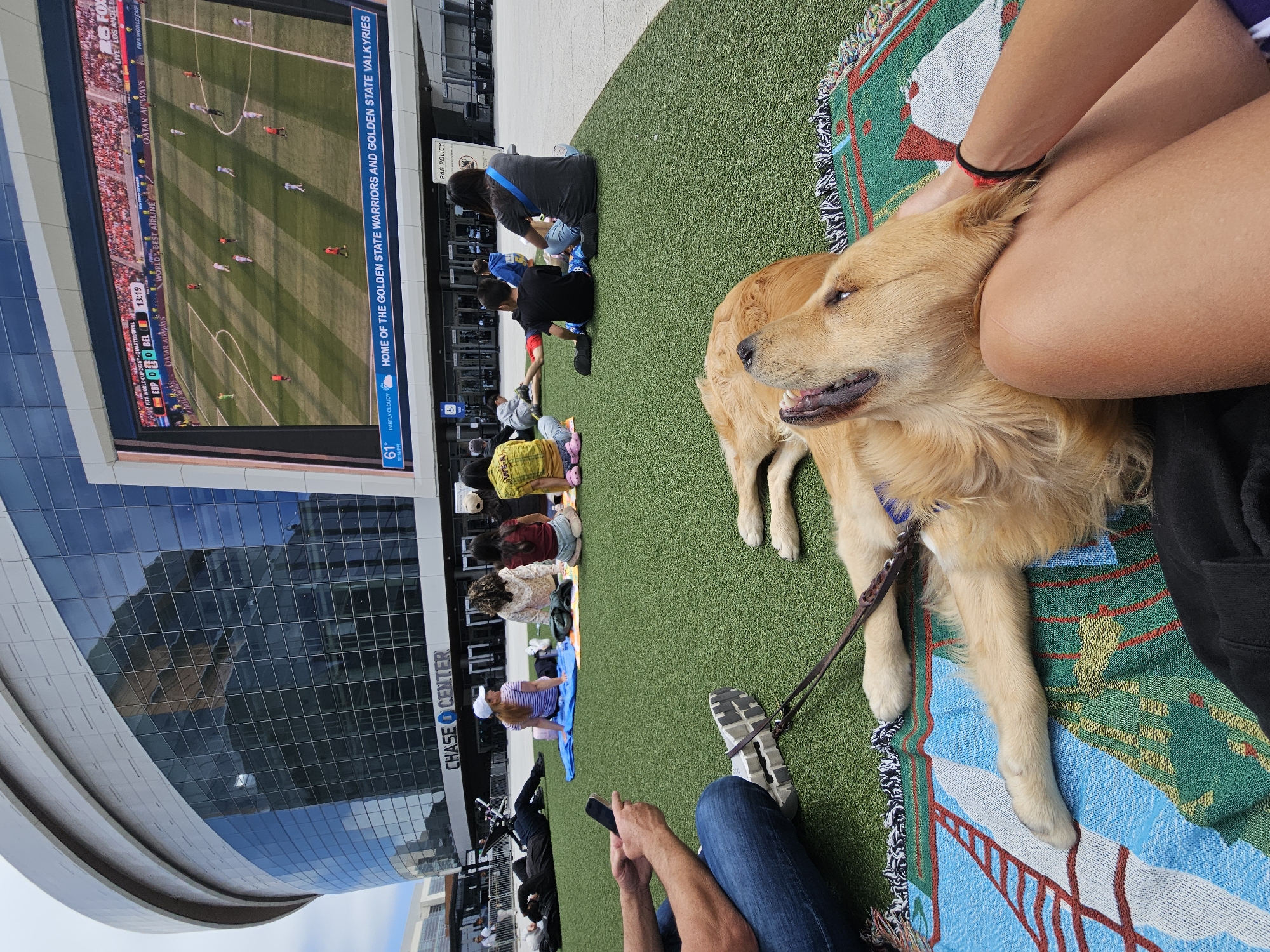 The image depicts an outdoor scene at the Chase Center in San Francisco, USA. A large screen displays a soccer game, with the score of Spain versus Belgium at 0-0. Below the screen, several people are seated on a green turf surface, some on blankets, facing the screen. In the foreground, a golden retriever is lying on a blanket, with its head resting on a person's lap. The person's legs and arm are visible. Other people are seated further back, some with their backs to the camera. The Chase Center building, with its distinctive architecture and glass facade, is visible in the background. The overall atmosphere appears relaxed and casual, with people enjoying the game outdoors.