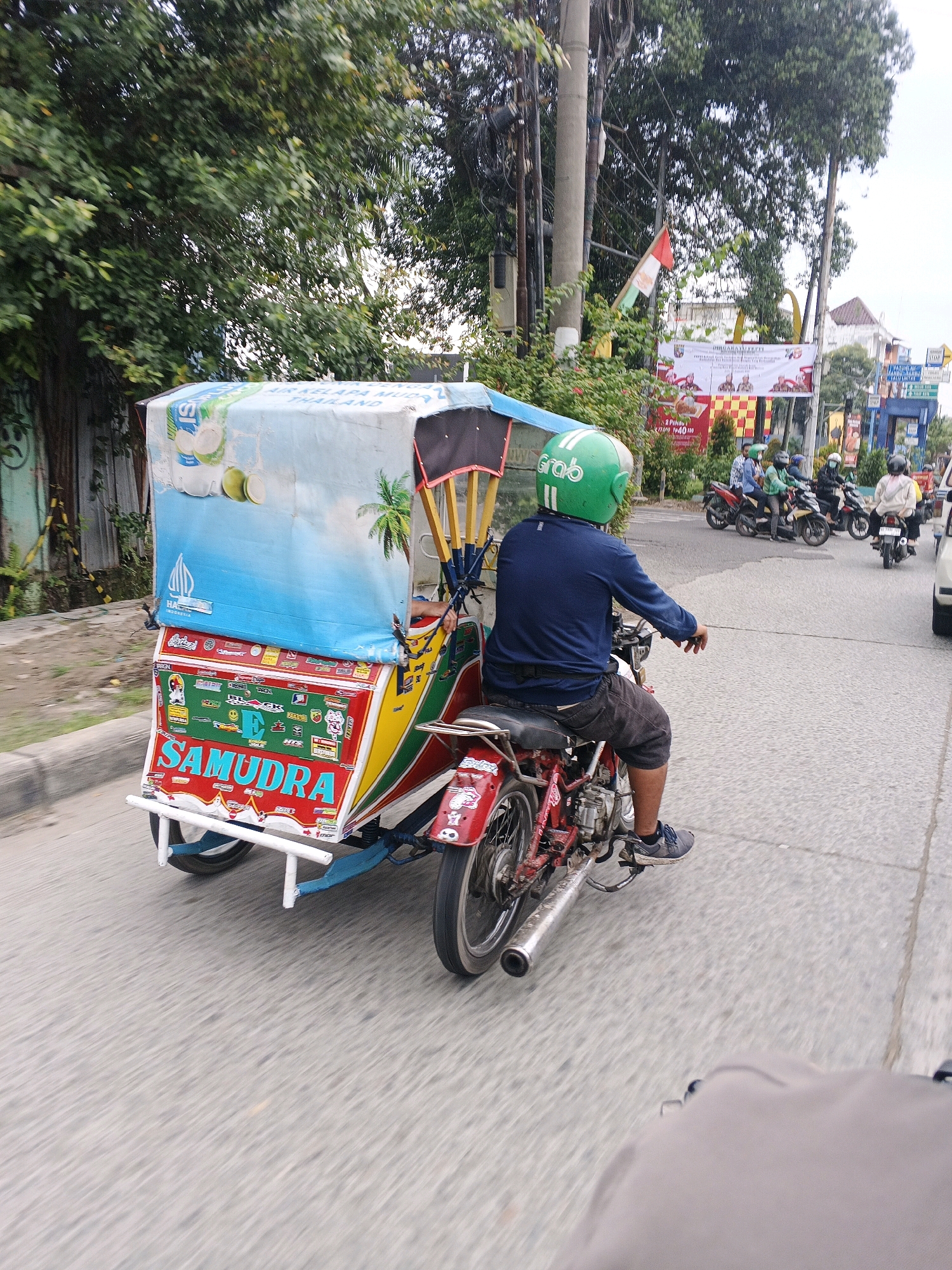 A side view captured from a low angle shows a colorful three-wheeled vehicle, likely a pedicab or becak, driving on an asphalt road in the City of Medan, Indonesia. The vehicle has a large canopy decorated with images of coconuts and a palm tree, along with the word "SAMUDRA" in large red letters. The driver, wearing a green helmet with a "Grab" logo, is seated on the motorcycle component of the vehicle, which is attached to the passenger cabin. The driver is wearing a dark blue long-sleeved shirt and dark shorts. Several other motorcyclists and their vehicles are visible in the background, also on the road. The environment includes trees lining the road and some buildings in the distance, with banners indicating commercial activity. The road surface is visible, with some blur suggesting motion.