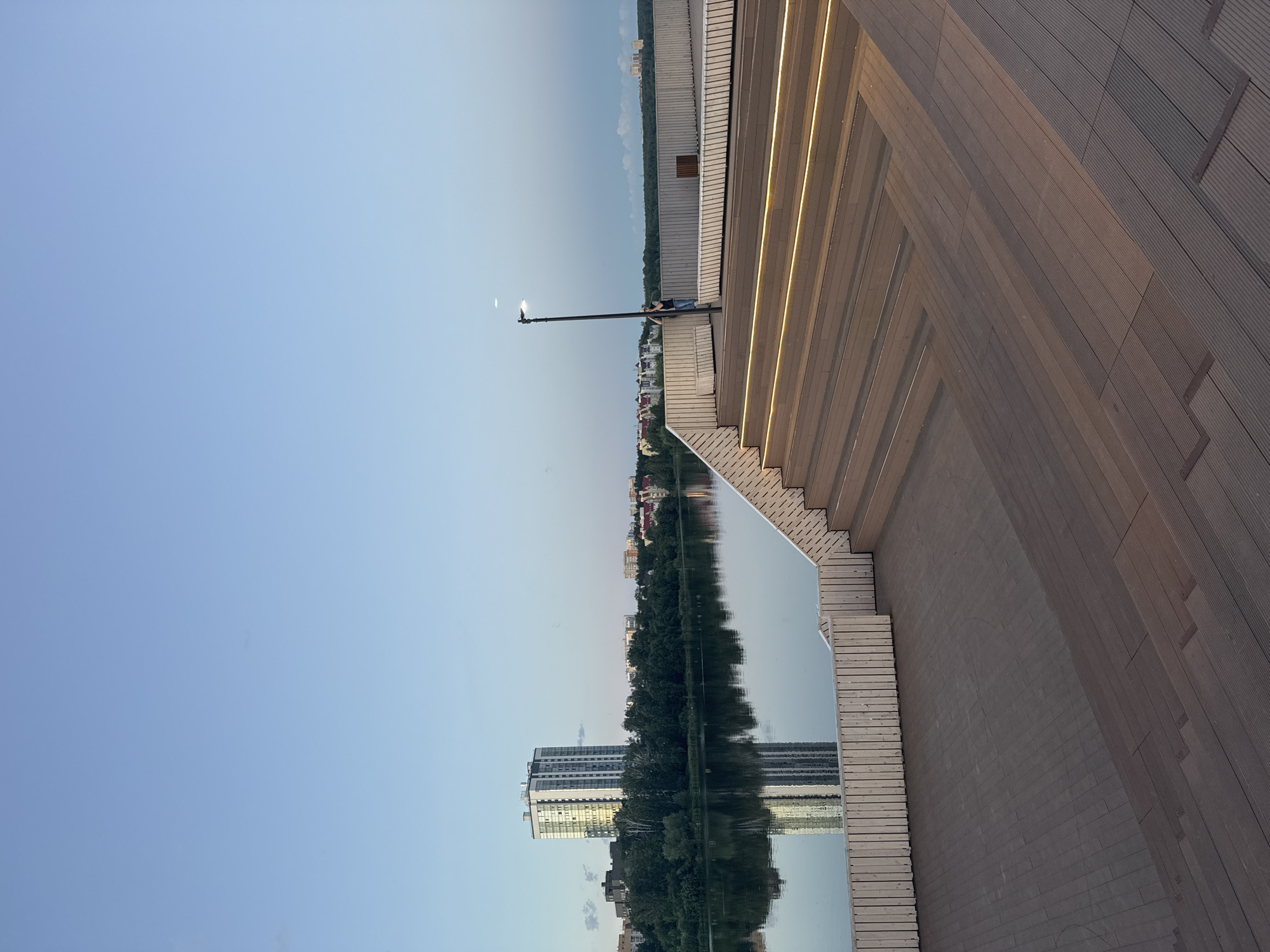 The image depicts the embankment of the Iset River in Yekaterinburg, Russia, during twilight. In the foreground, a modern wooden amphitheater with integrated lighting rises from the river. Beyond the amphitheater, the calm river reflects the sky and the cityscape. Across the river, a dense line of green trees separates the water from a row of residential buildings and a prominent tall, white apartment building. The sky is clear and pale blue, with a hint of lighter tones near the horizon, suggesting the transition from day to night. A single streetlamp stands on the embankment, and a person can be seen in the distance near it. The overall scene conveys a sense of urban tranquility and modern public space design.