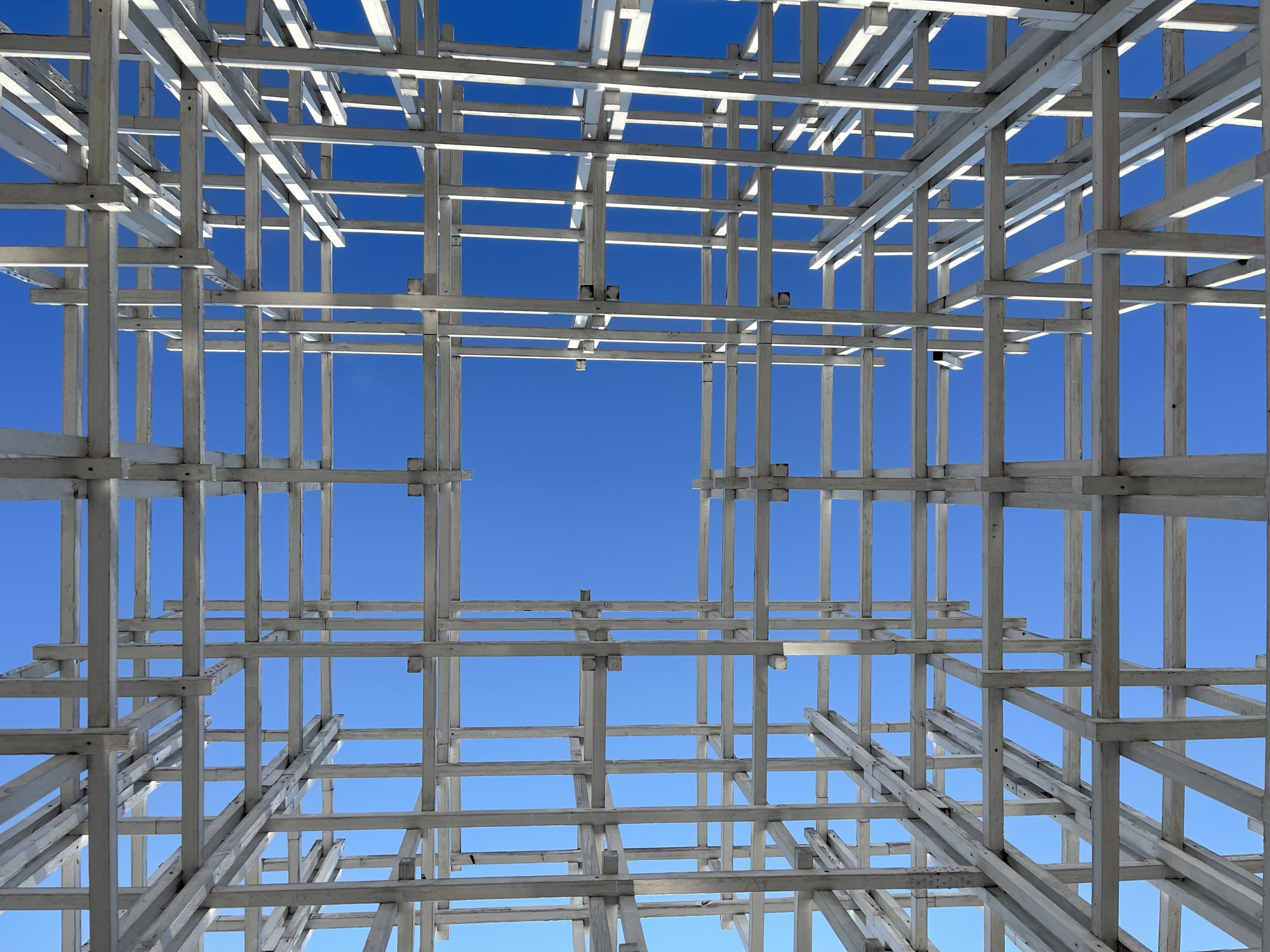 The image shows a perspective view looking upwards through a white wooden lattice structure against a bright blue sky. The lattice is composed of numerous horizontal and vertical beams intersecting at various points, creating a grid-like pattern with open squares and rectangles. The construction appears to be unfinished or a design element of a building or outdoor space. There are no people, animals, or other discernable objects in the image besides the wooden structure and the sky. The lighting suggests it is daytime, with the sun casting some subtle shadows on the beams. The location is not specified within the image itself.