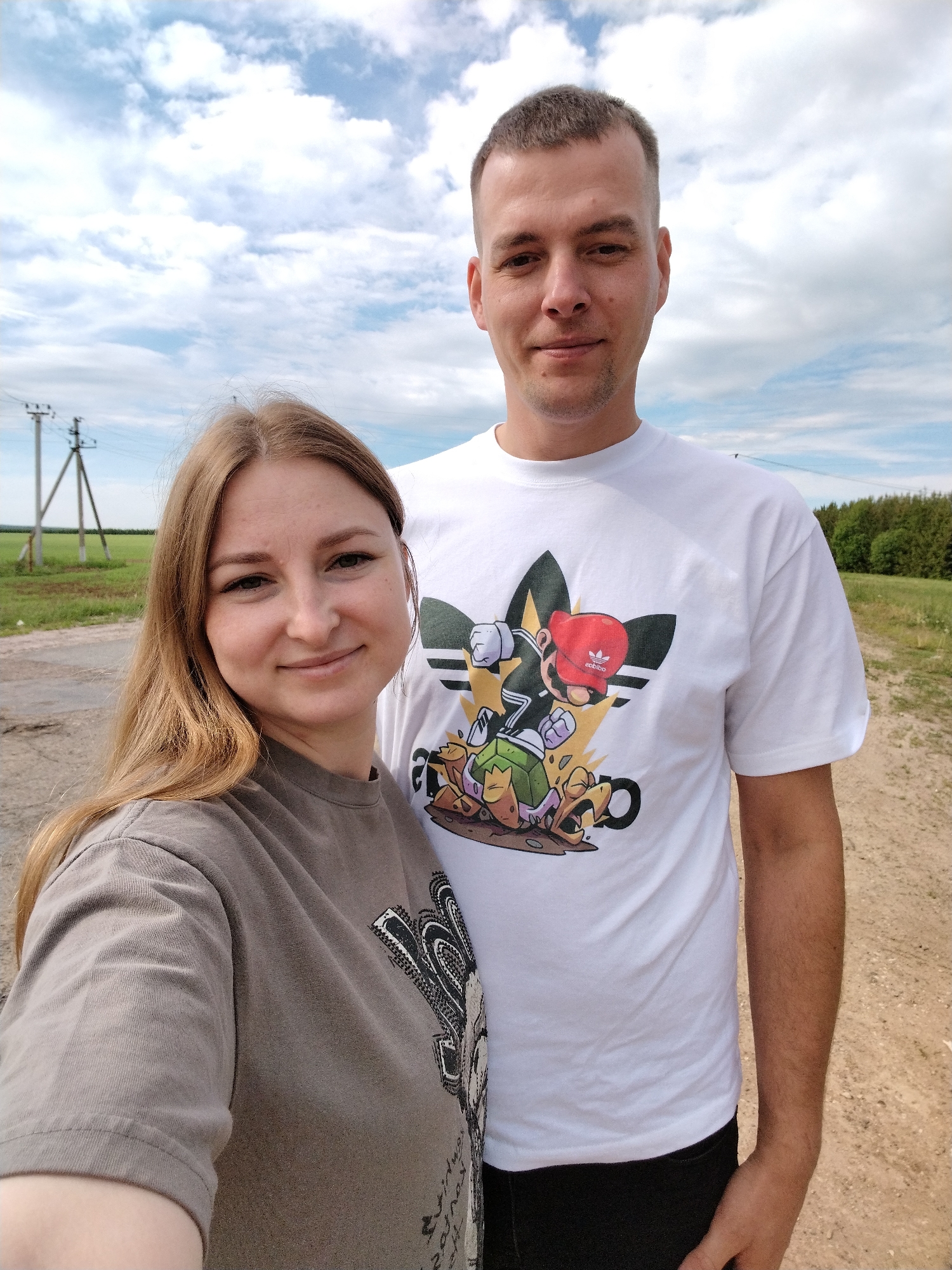 A digital photograph depicts a fair-skinned woman and man standing outdoors in a rural setting. The woman, on the left, has long, light brown hair and wears a heathered gray t-shirt with a stylized design. She is holding the camera and smiling slightly. The man, standing to her right and slightly behind her, is taller with short, light brown hair and a white t-shirt featuring a graphic combining the Adidas logo with Mario Brothers characters.
The background includes a dirt path or road, grassy fields, and a line of trees in the distance. Electrical poles are visible in the left background. The sky is partially cloudy with a mix of blue and white. The photograph was taken in a rural region near Tavlarovo, Russia.